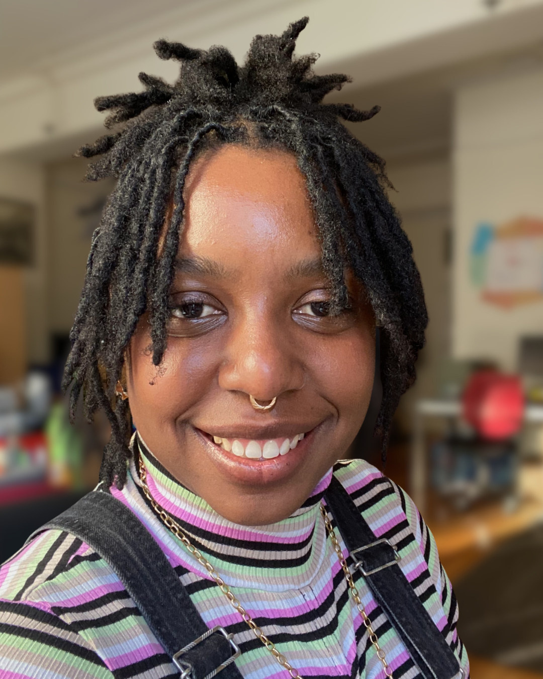 A woman with short curly dark hair, wearing a dark sleeveless top, smiling against a gray background.