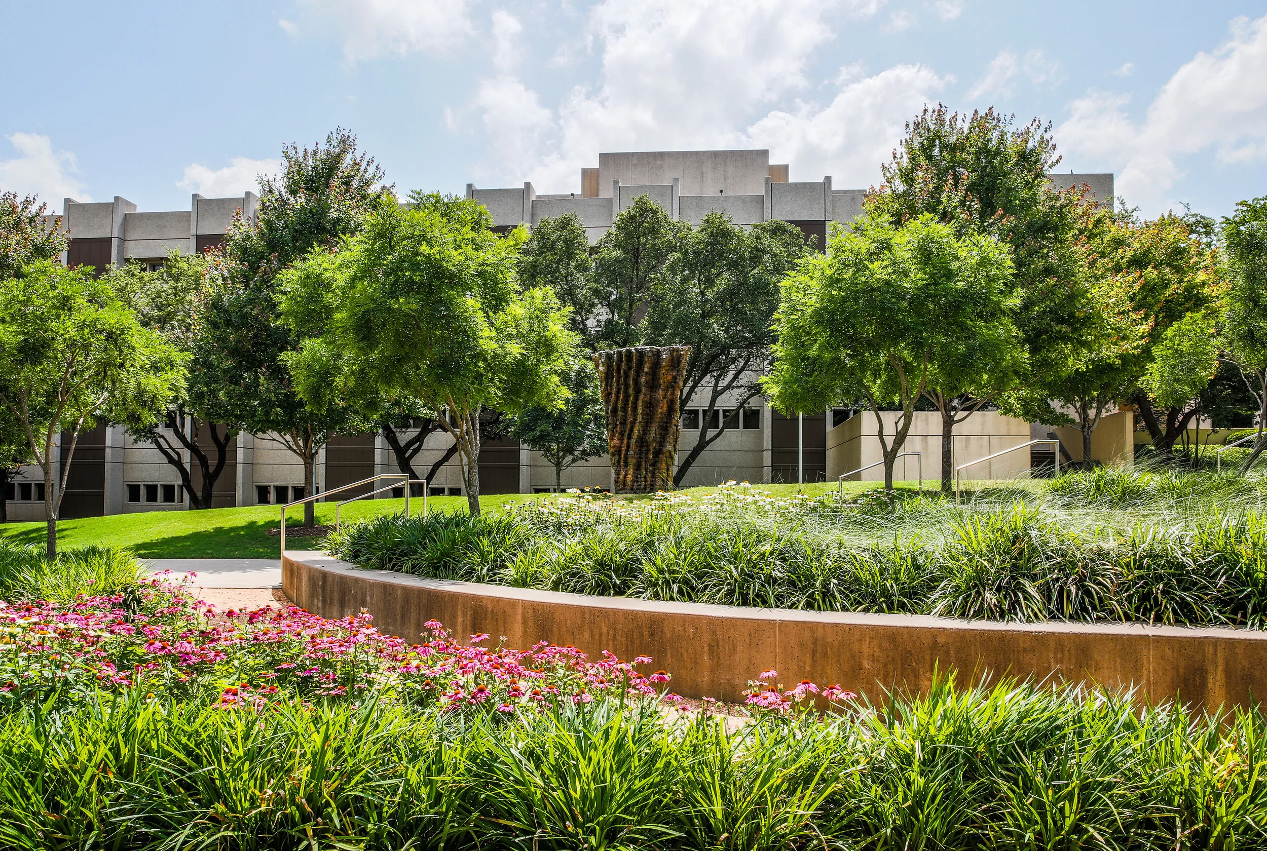 Lush green park with trees, flowers, and a concrete path leading to a modern building in the background.