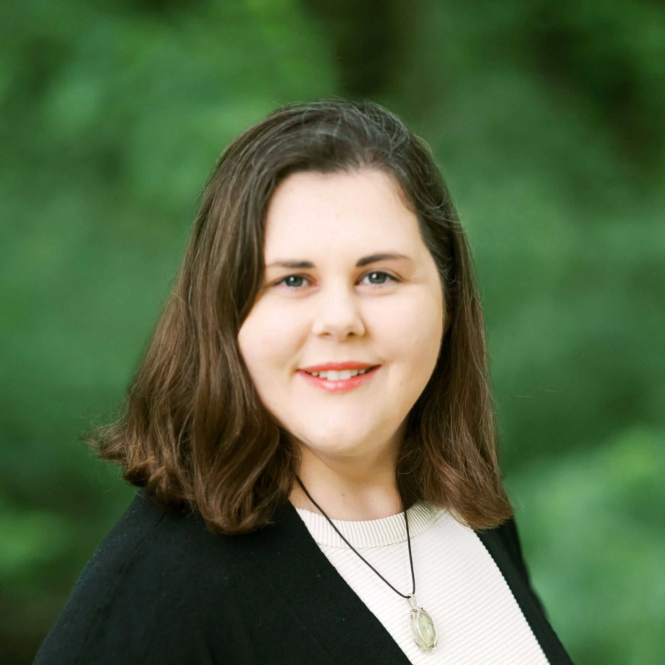 Woman with brown hair wearing a black sweater and pendant necklace, smiling with greenery in the background.