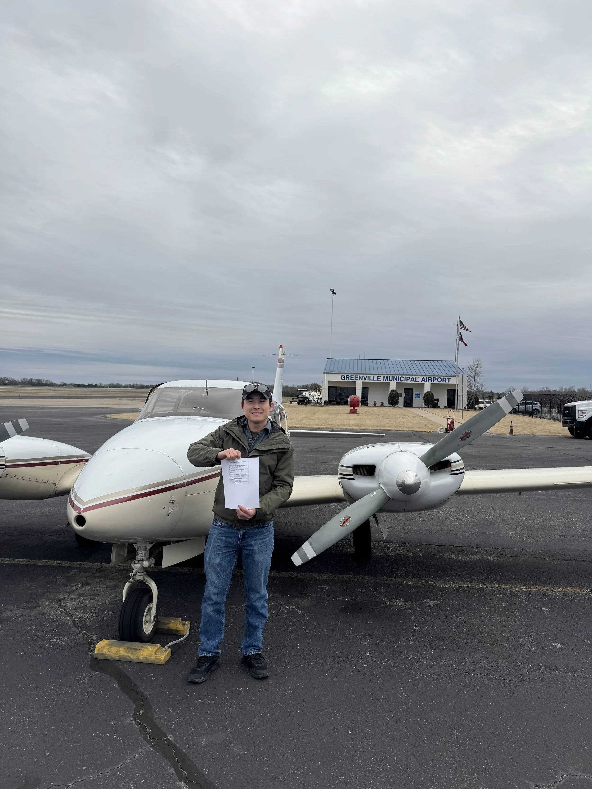 A young man stands next to a small white airplane at Greenville Municipal Airport, holding a piece of paper, with a building in the background.