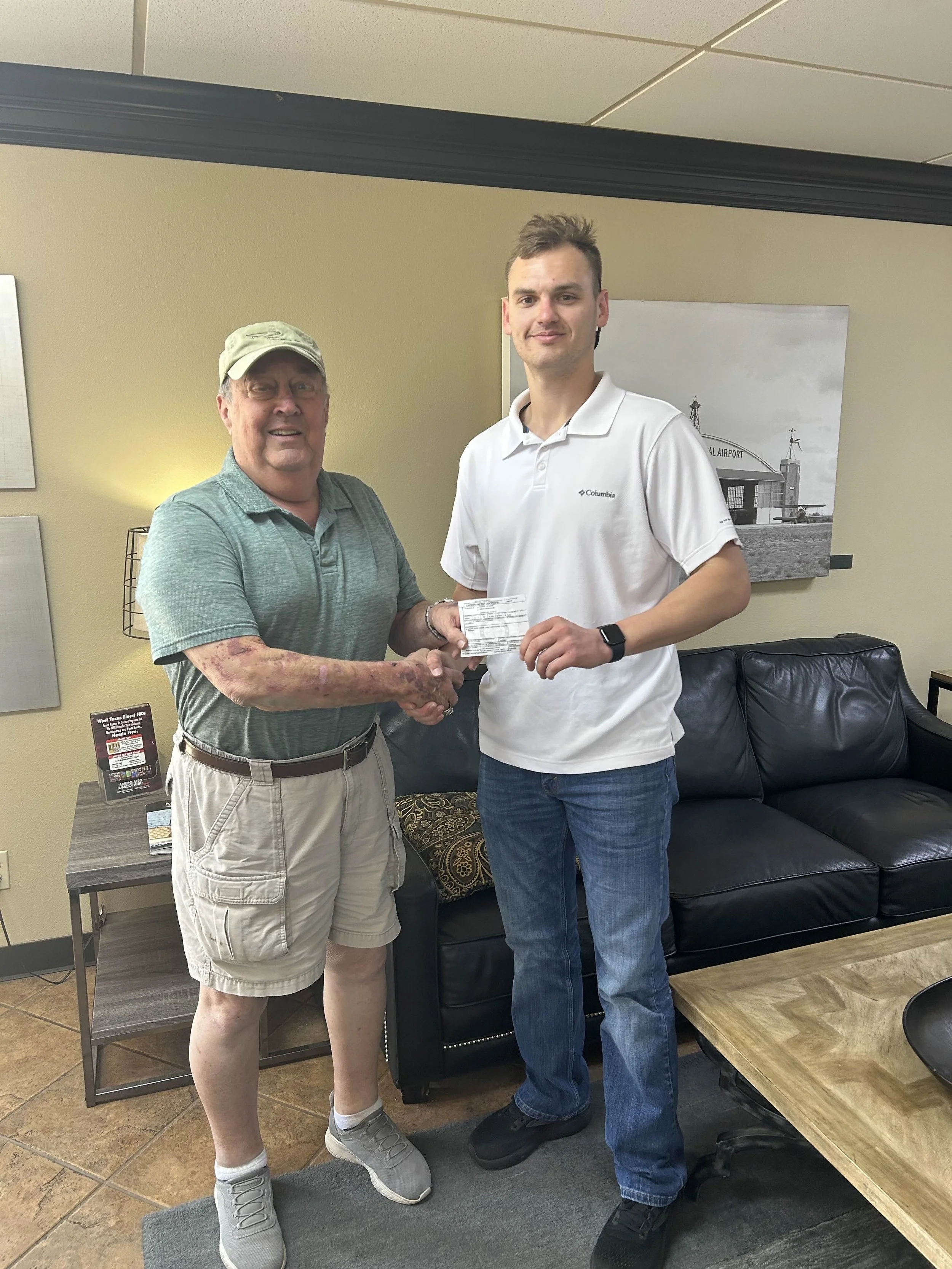 A man in casual clothing shaking hands with a young man in a white polo shirt, holding a document, in a waiting room with a black leather couch and artwork on the wall.