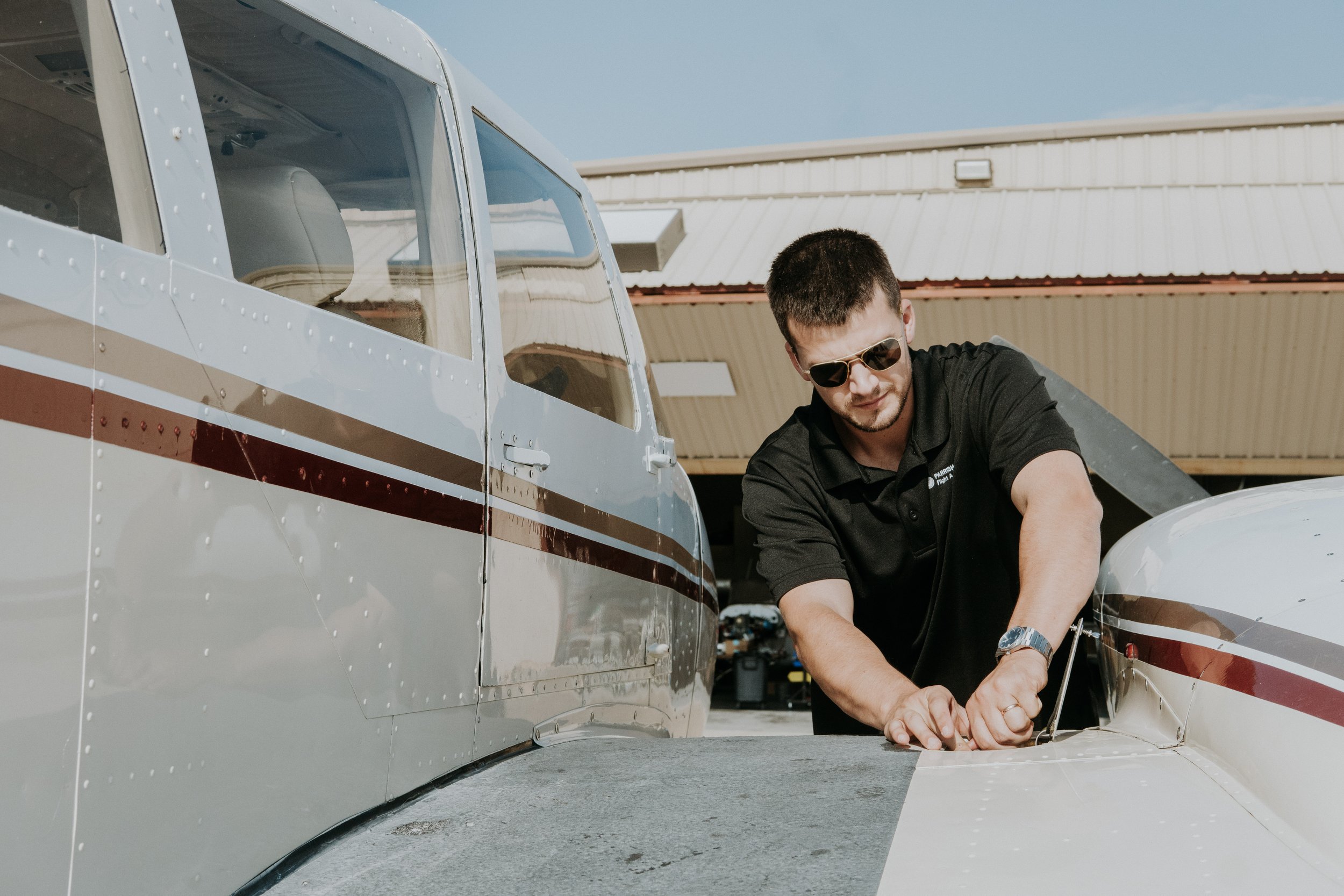 A man wearing sunglasses and a black polo shirt working on the wing of a small airplane outside a hangar.