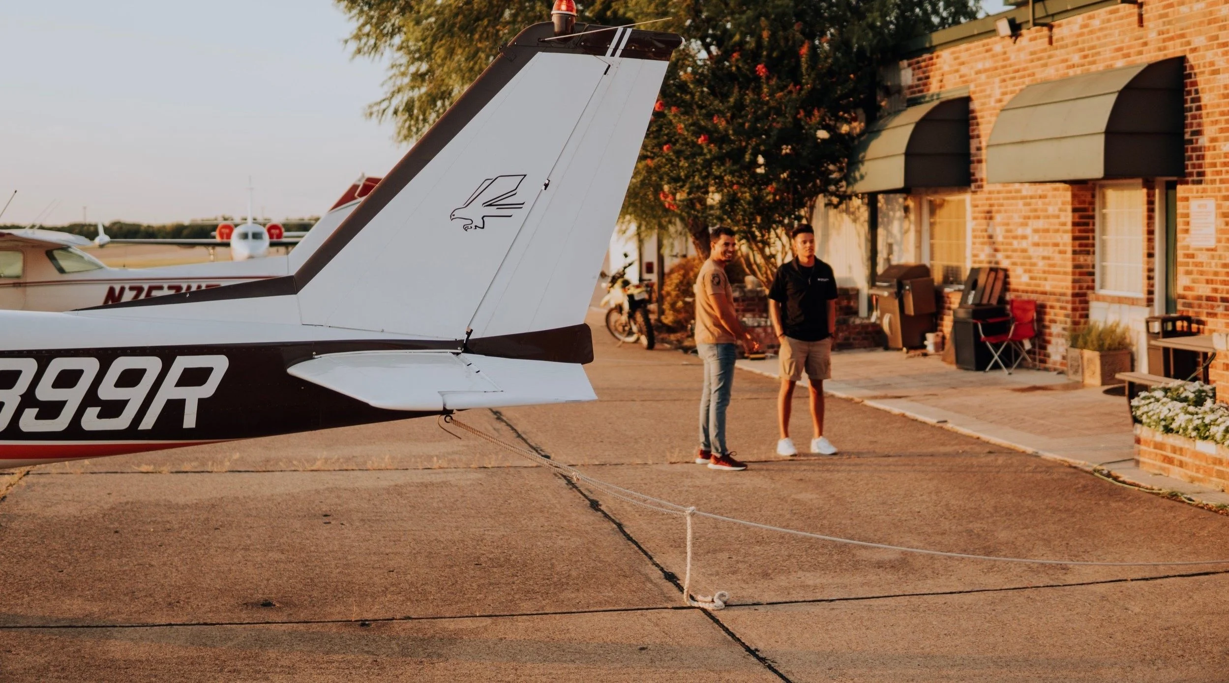 Two young men standing near a small airplane parked on a tarmac in front of a brick building with green awnings, during sunset.
