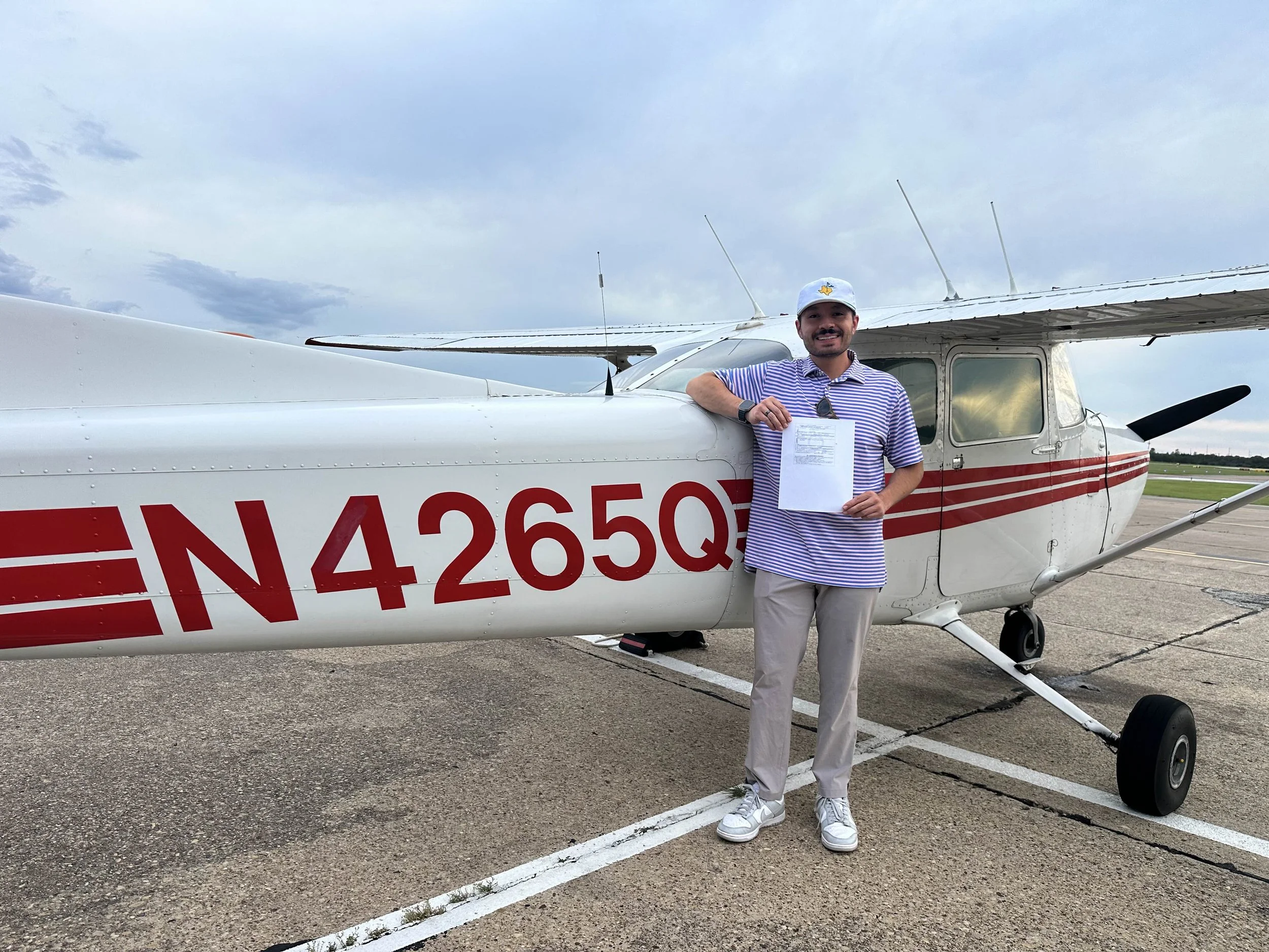 A man standing next to a small white airplane with red stripes, holding a paper and smiling. He is wearing a striped polo shirt, beige pants, white sneakers, a watch, sunglasses hanging from his shirt, and a white cap. The airplane is parked on an ai