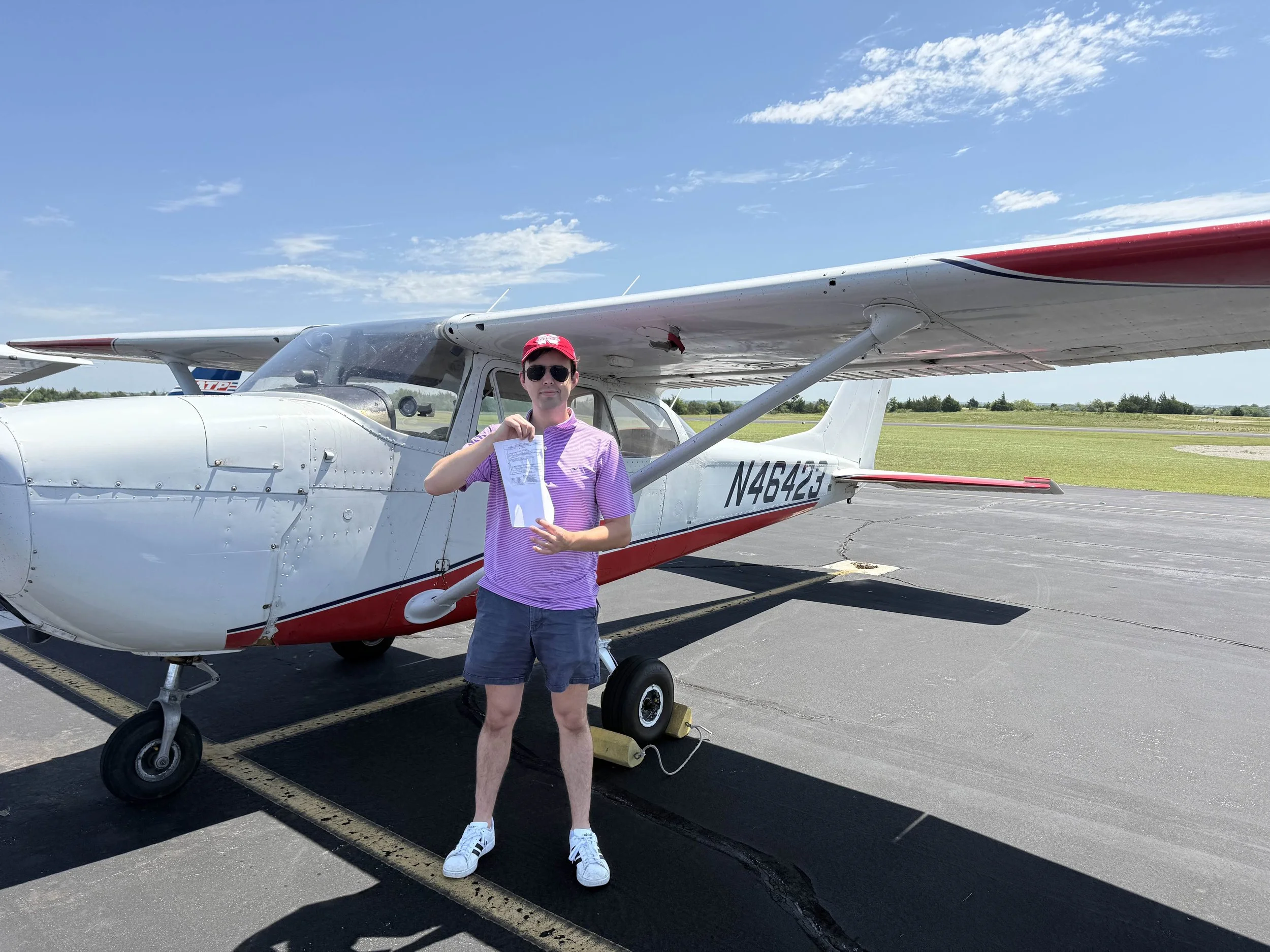 A young man in a purple shirt, shorts, white shoes, sunglasses, and a red cap holding a document stands in front of a small white and red airplane parked on an airport tarmac, with grass and blue sky in the background.