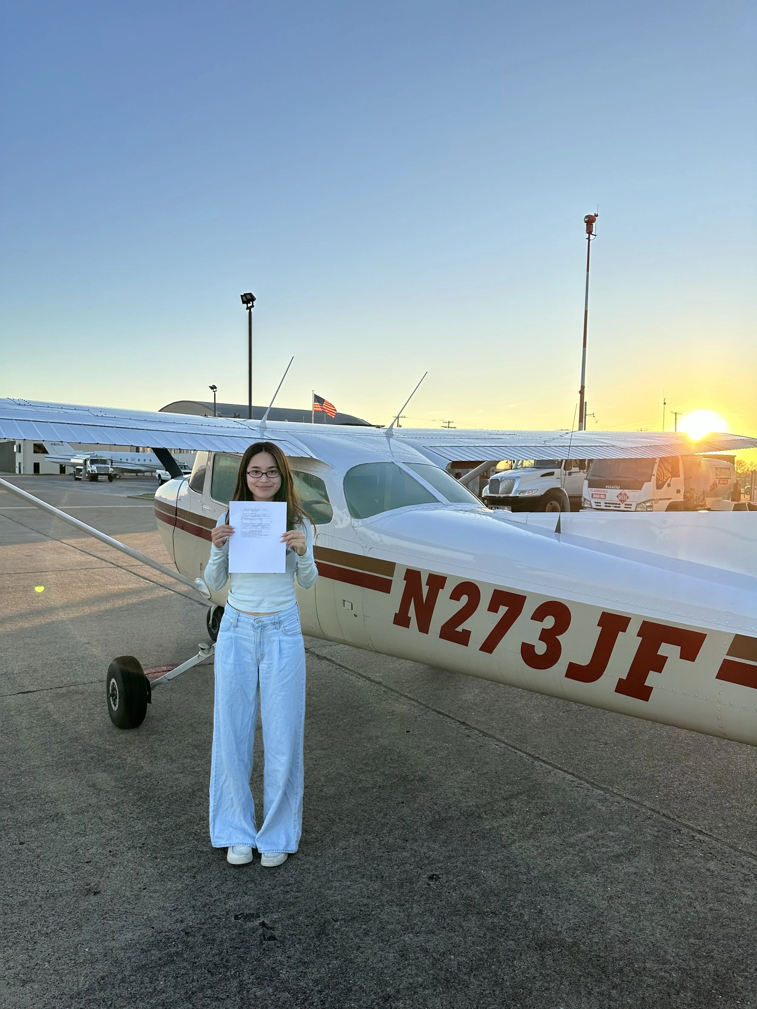 A young woman in light blue pants and a white long-sleeve shirt standing in front of a small white airplane with red markings, holding a piece of paper, at an airport during sunset.