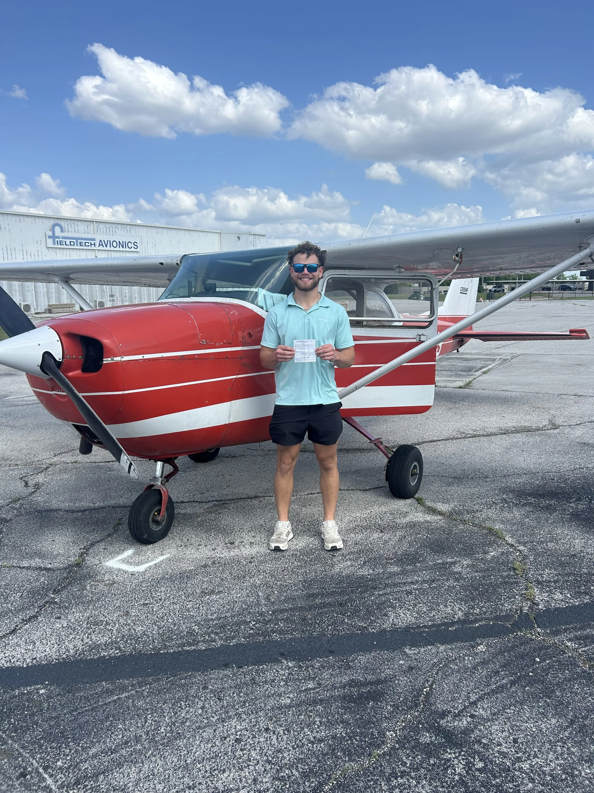 A smiling man wearing sunglasses, a light blue polo shirt, black shorts, and sneakers holding a paper standing in front of a small red and white airplane on an airport tarmac with blue skies and clouds overhead.