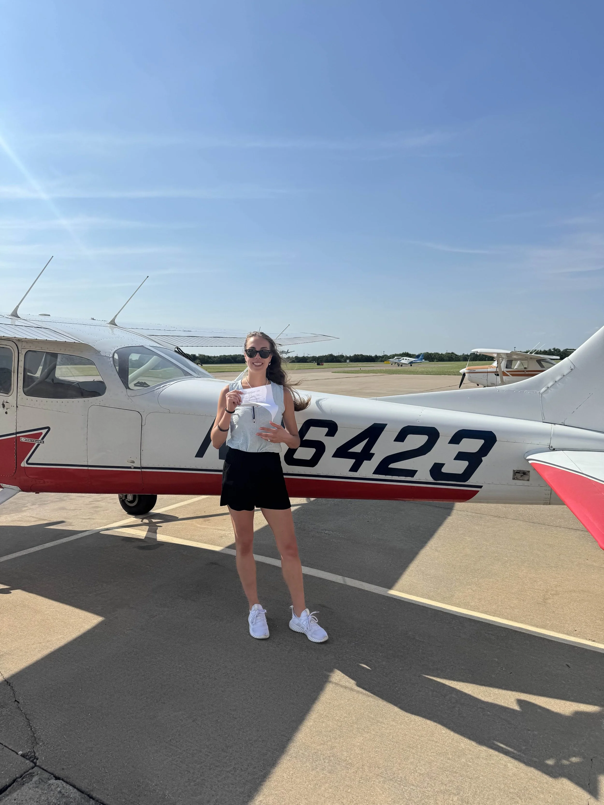 A young woman stands on the tarmac holding a piece of paper or boarding pass while smiling in front of a small airplane with the registration number 164423. She is wearing sunglasses, a sleeveless shirt, black shorts, and white sneakers, with a clear
