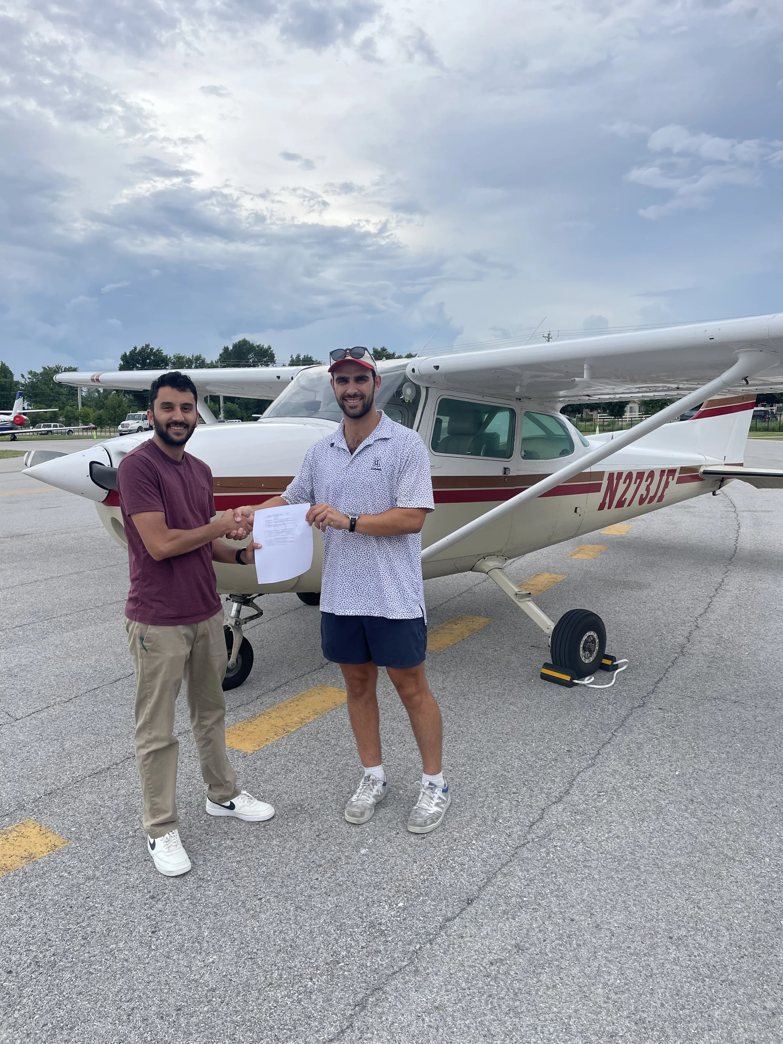 Two men shaking hands in front of a small airplane, with one man holding a document, on an airport tarmac under a cloudy sky.