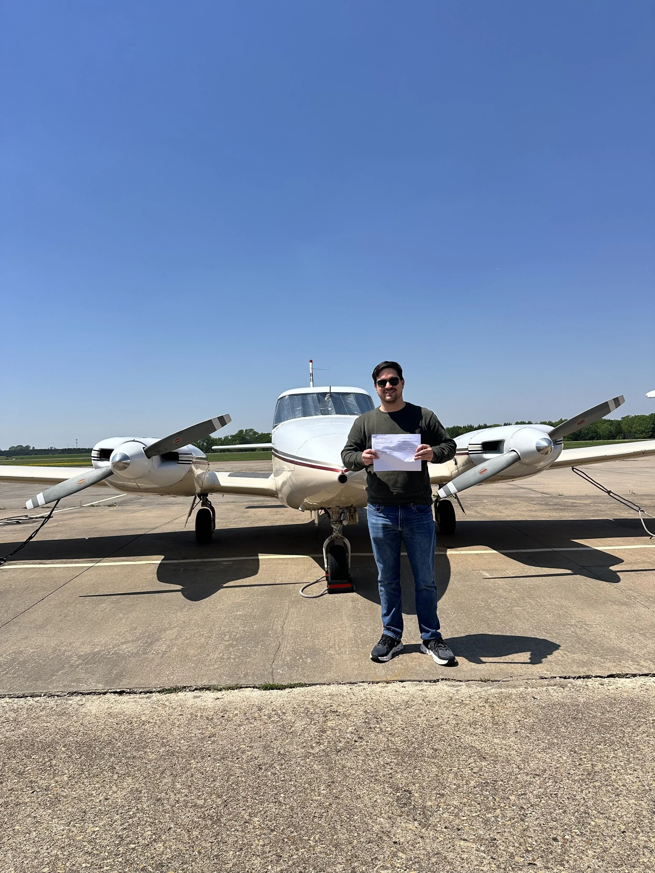 Man standing in front of a twin-engine airplane on an airstrip, holding a piece of paper and wearing sunglasses.
