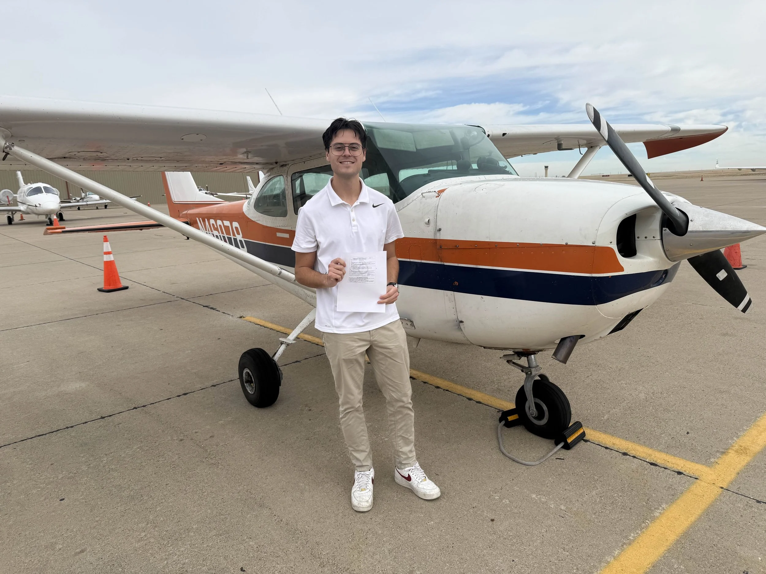 Young man holding a paper standing in front of a small propeller airplane at an airport, with other small planes in the background and orange traffic cones on the tarmac.