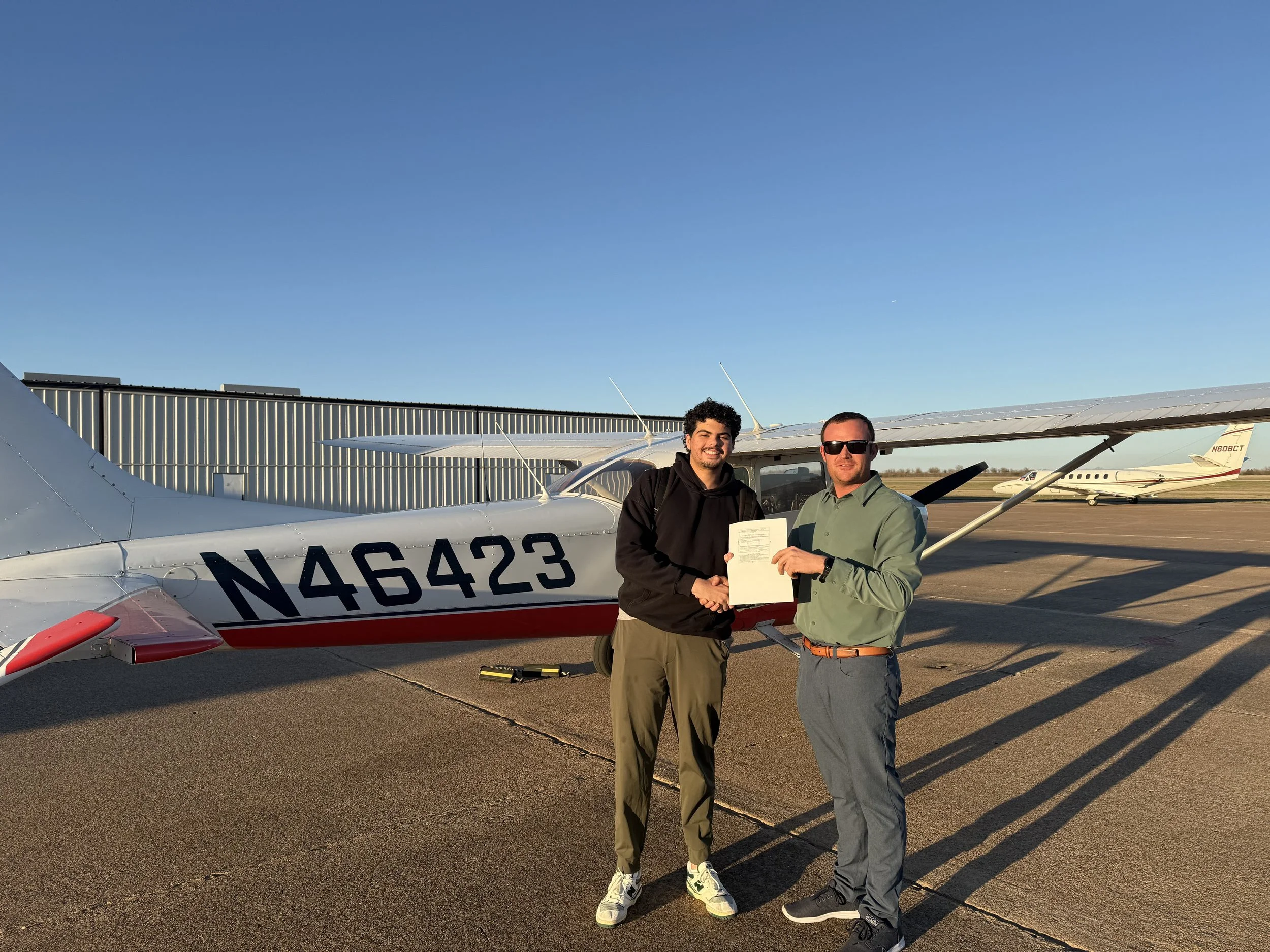 Two men shake hands on airport tarmac in front of a small white airplane with red accents, one of them holding a document, with another plane and hangar in the background during sunset.