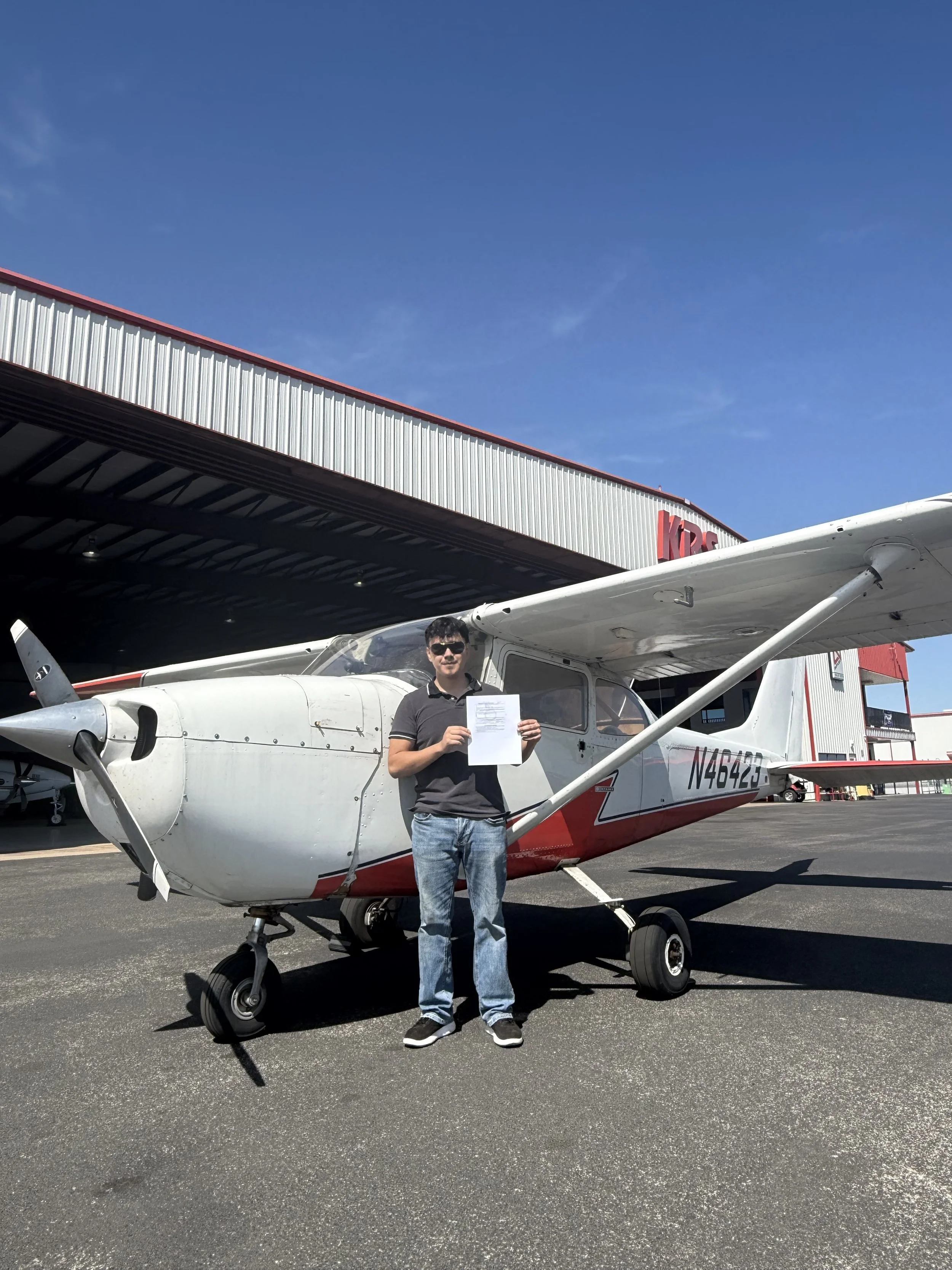 Man standing in front of a small airplane holding a signed document outside an aviation hangar on a clear day.