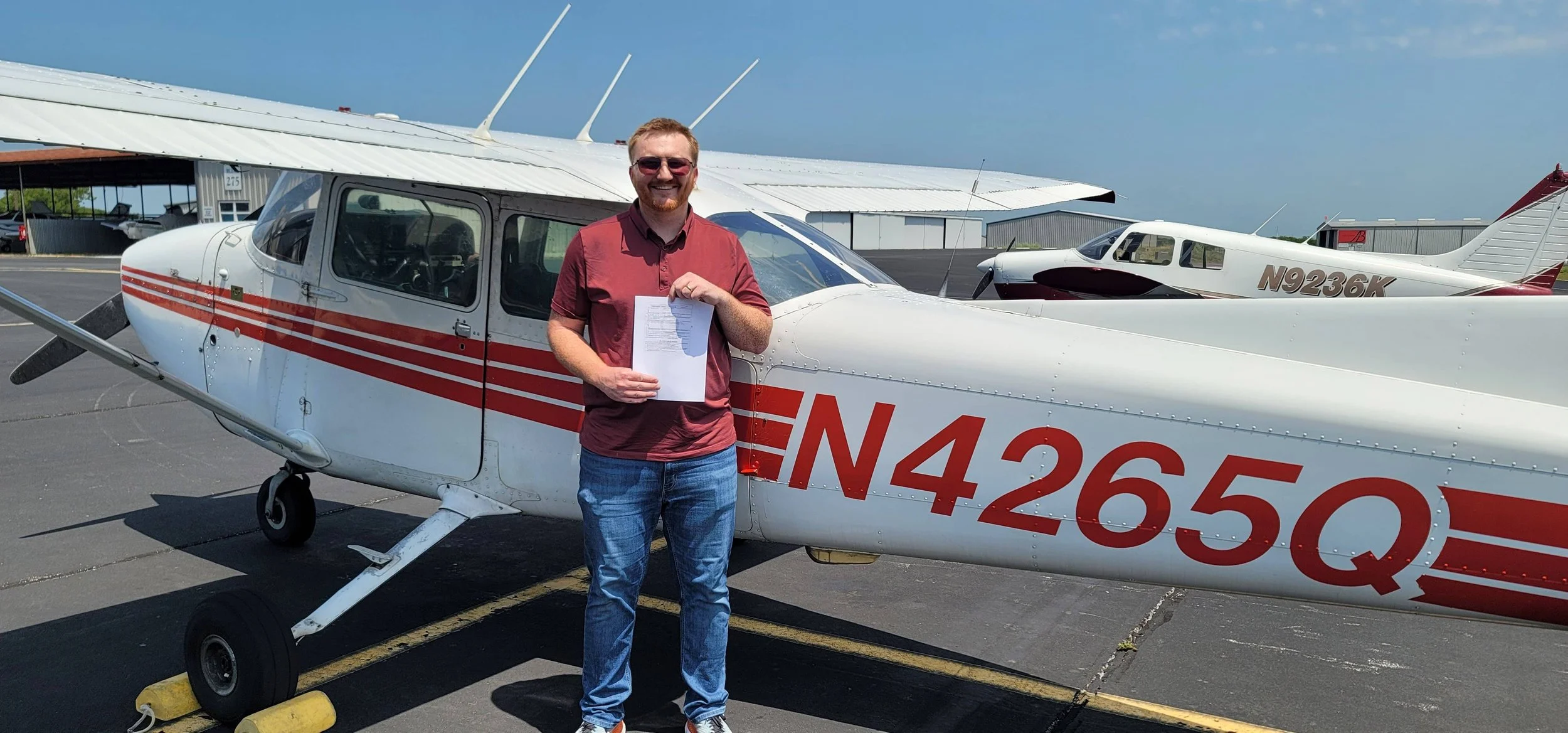 Man standing next to a small white propeller airplane with red stripes, holding a piece of paper. The airplane has the registration number N4265Q. The scene is at an airport or airfield on a sunny day.