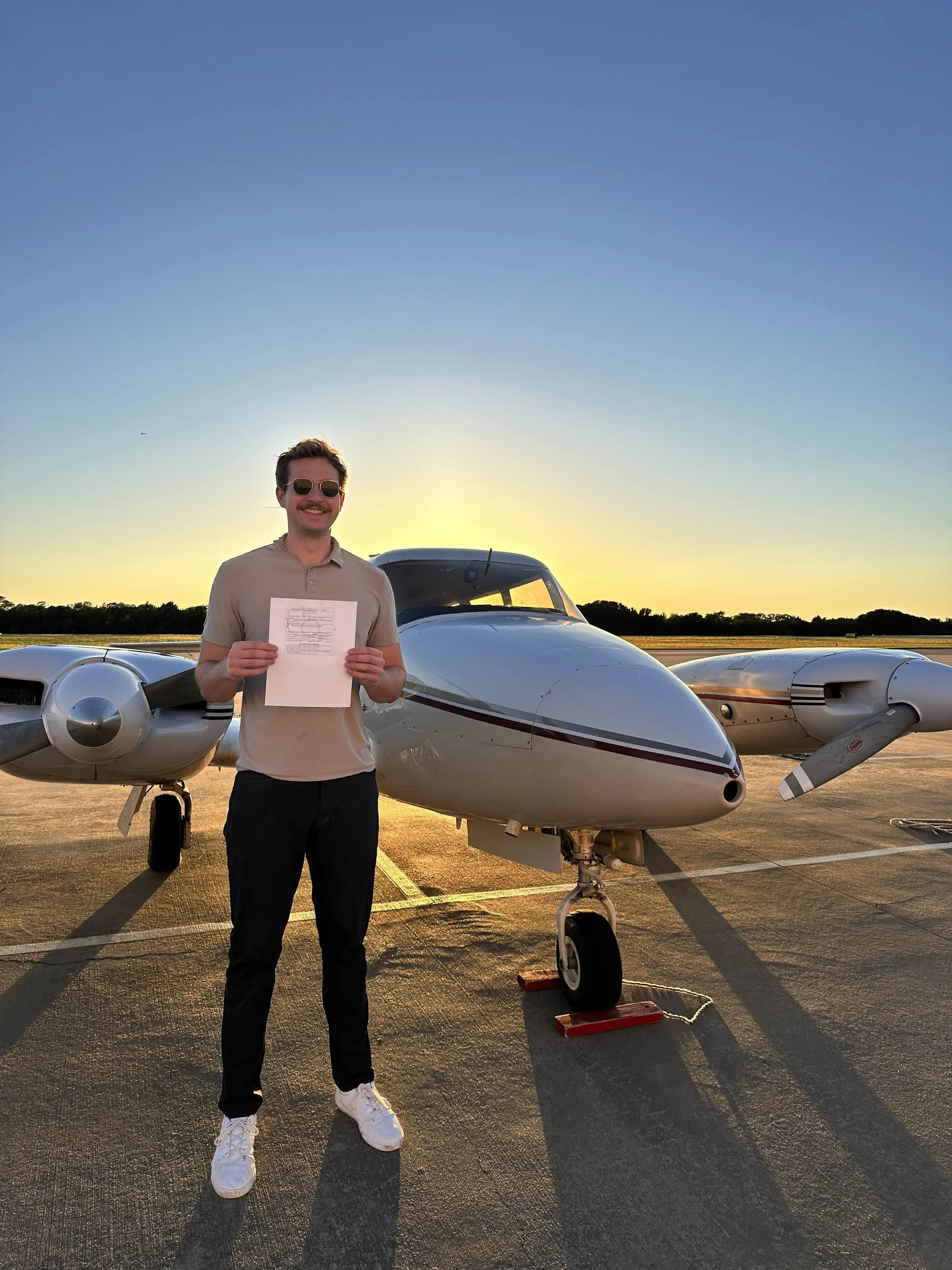Man with sunglasses and a paper standing in front of a small airplane on an airport tarmac at sunset.