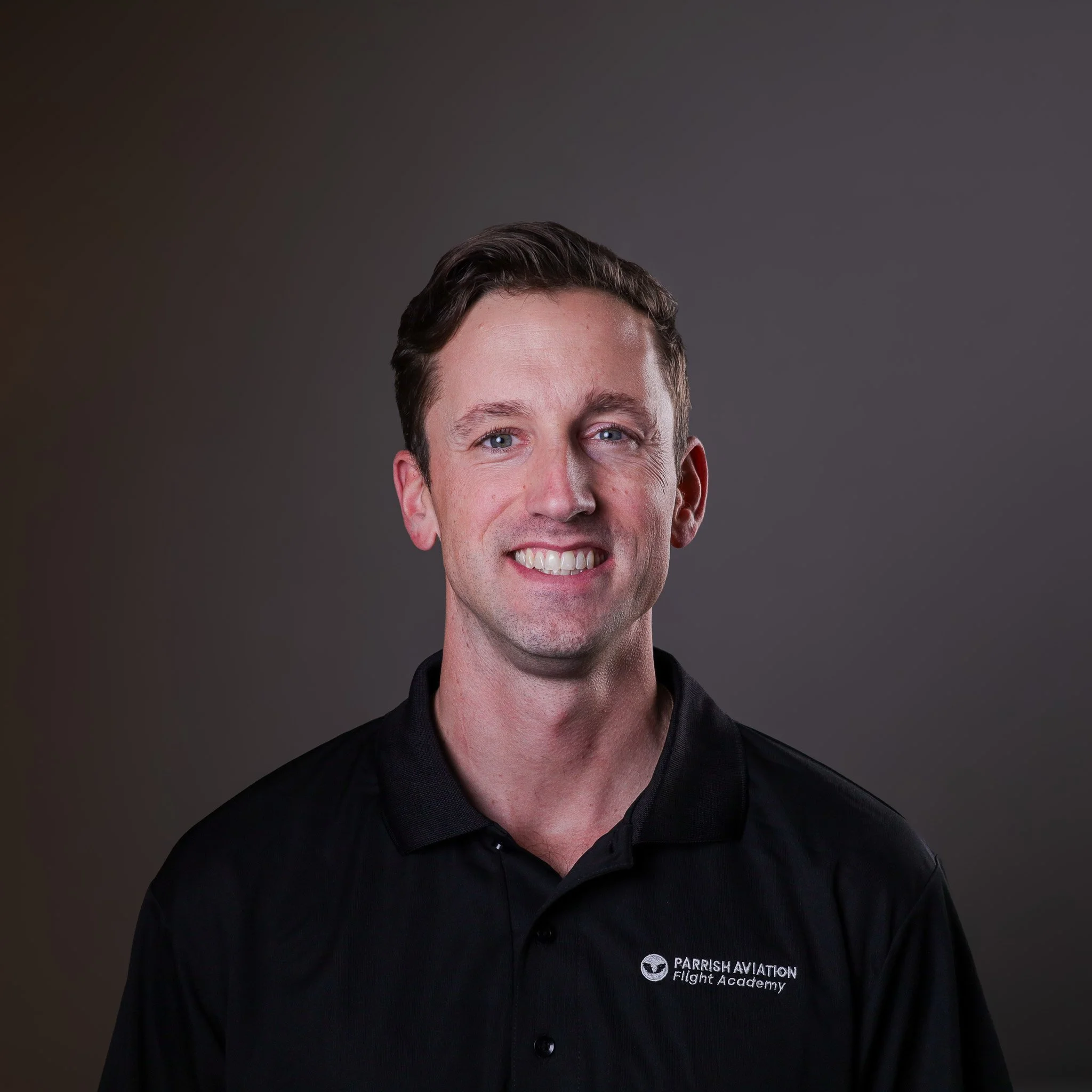 Portrait of a man in a black polo shirt with 'Parish Aviation Flight Academy' logo, smiling against a dark gradient background.