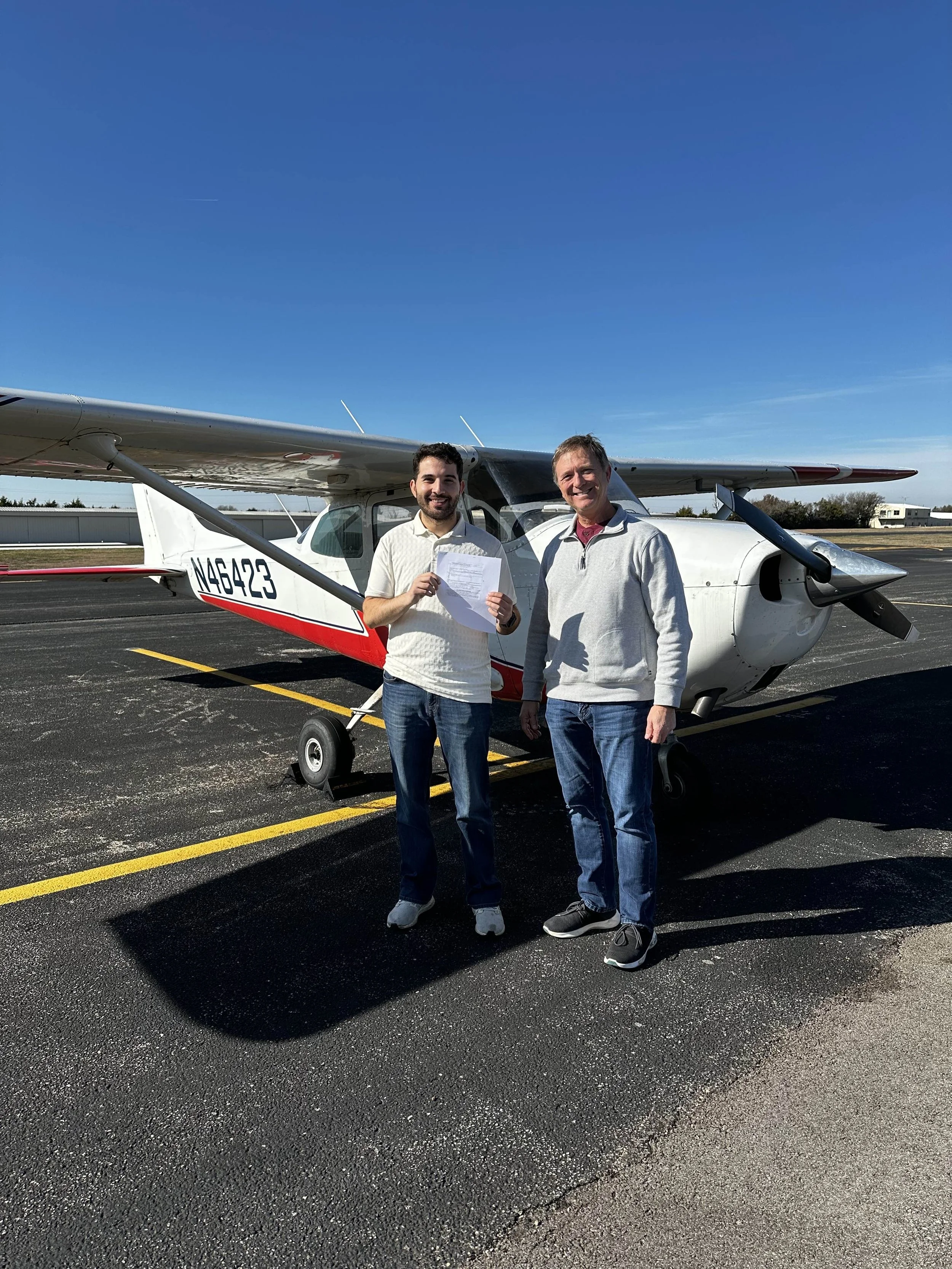 Two men standing in front of a small airplane on a tarmac, smiling, one holding a document, clear blue sky.