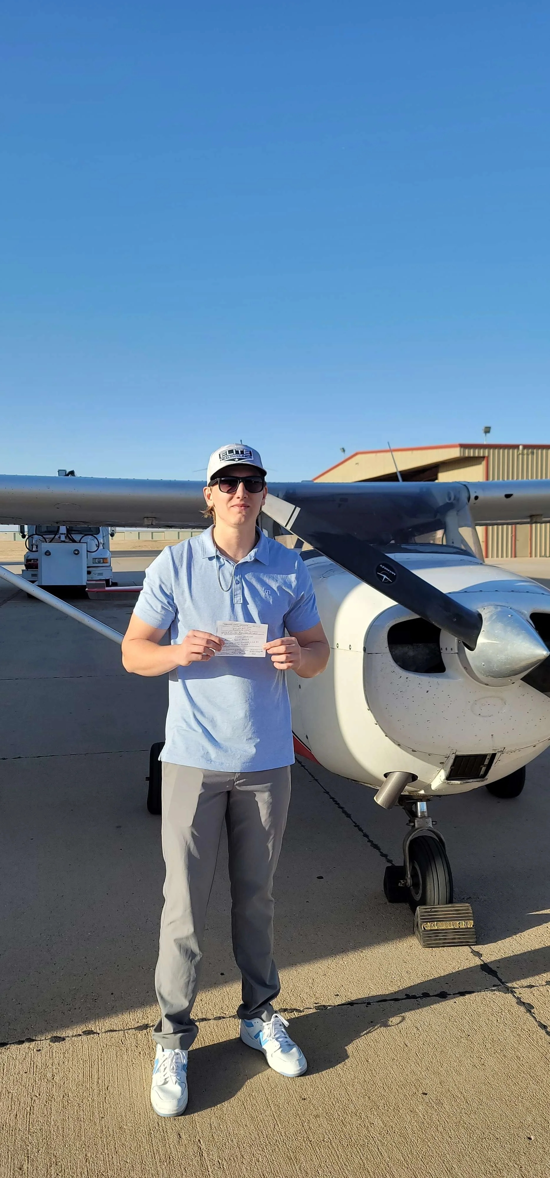 A young man wearing sunglasses, a gray cap, a light blue polo shirt, and gray pants standing on an airport tarmac in front of a small aircraft, holding a document or ticket. The plane is white with black propeller blades, and hangars are visible in t
