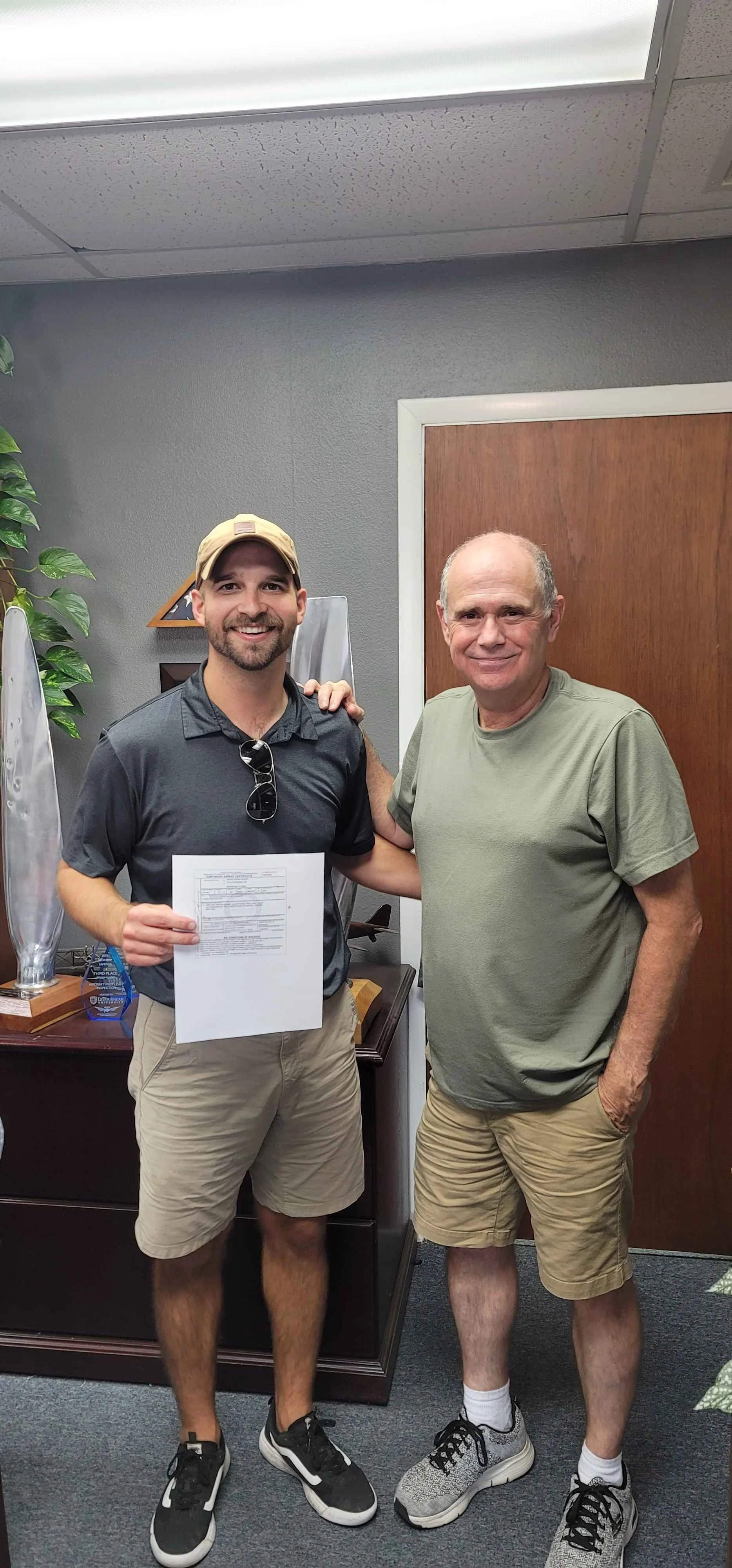 Two men standing together in an office, smiling at the camera. The younger man on the left is holding a document and has sunglasses hanging from his shirt collar. The older man on the right has his arm around the younger man's shoulder, wearing a gre