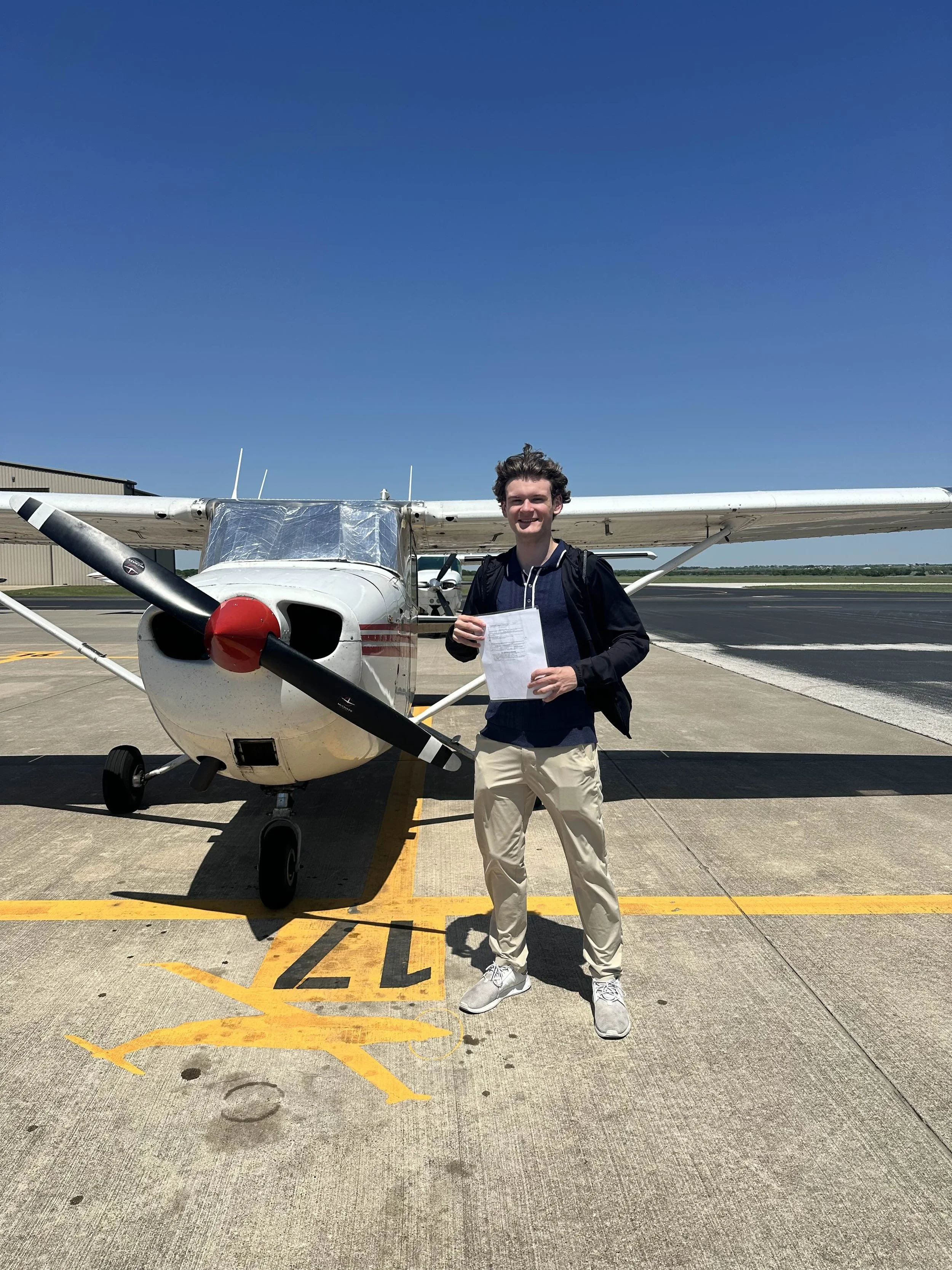 Young man holding a paper standing in front of a small airplane on the tarmac.