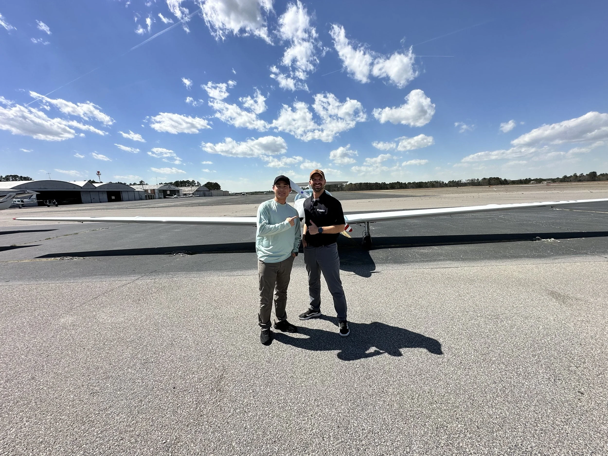 Two men smiling and standing in front of a small aircraft on an airfield, with hangars and a partly cloudy sky in the background.