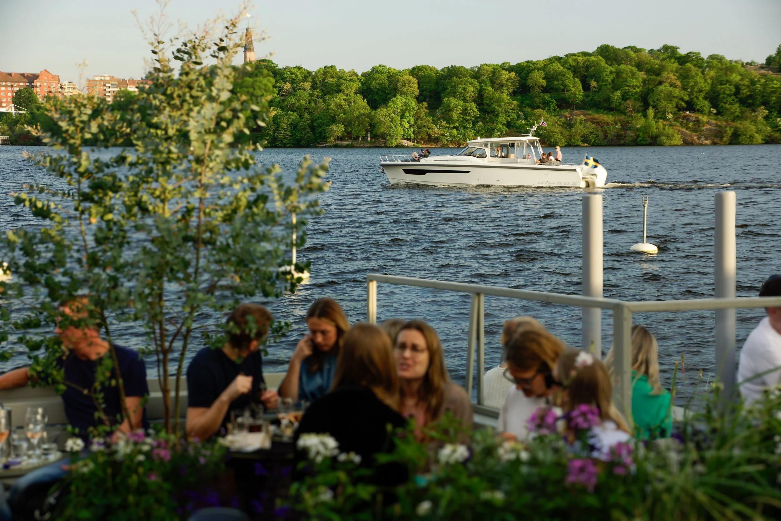Fokusera på en båt som flyter på Mälaren med suddiga människor som sitter vid Mälarpaviljongen bland blommorna en solig eftermiddag i Stockholm.