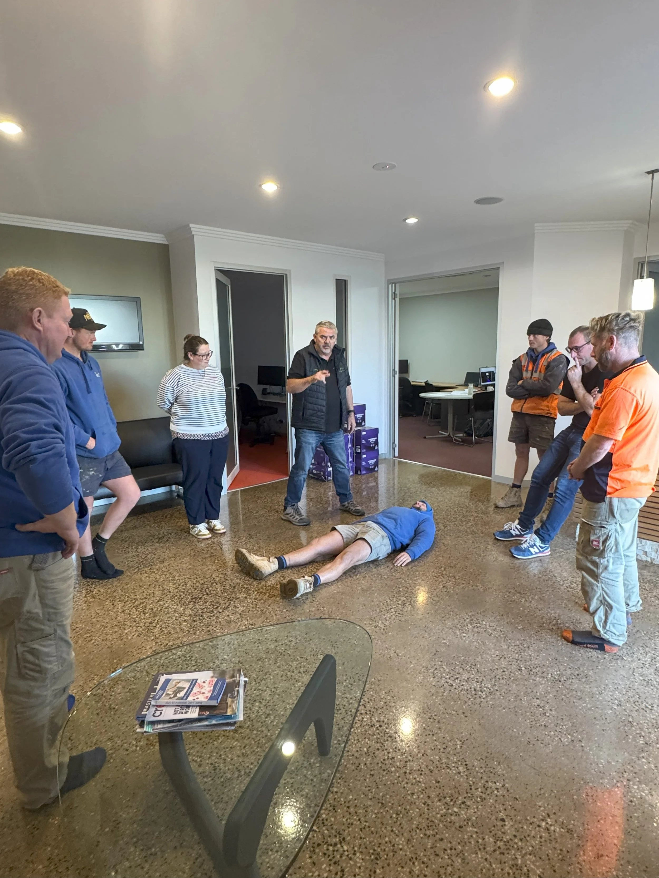 A person lies on the floor while a group of eight people stand around showing attention, possibly participating in a CPR or first aid training session in an office setting.
