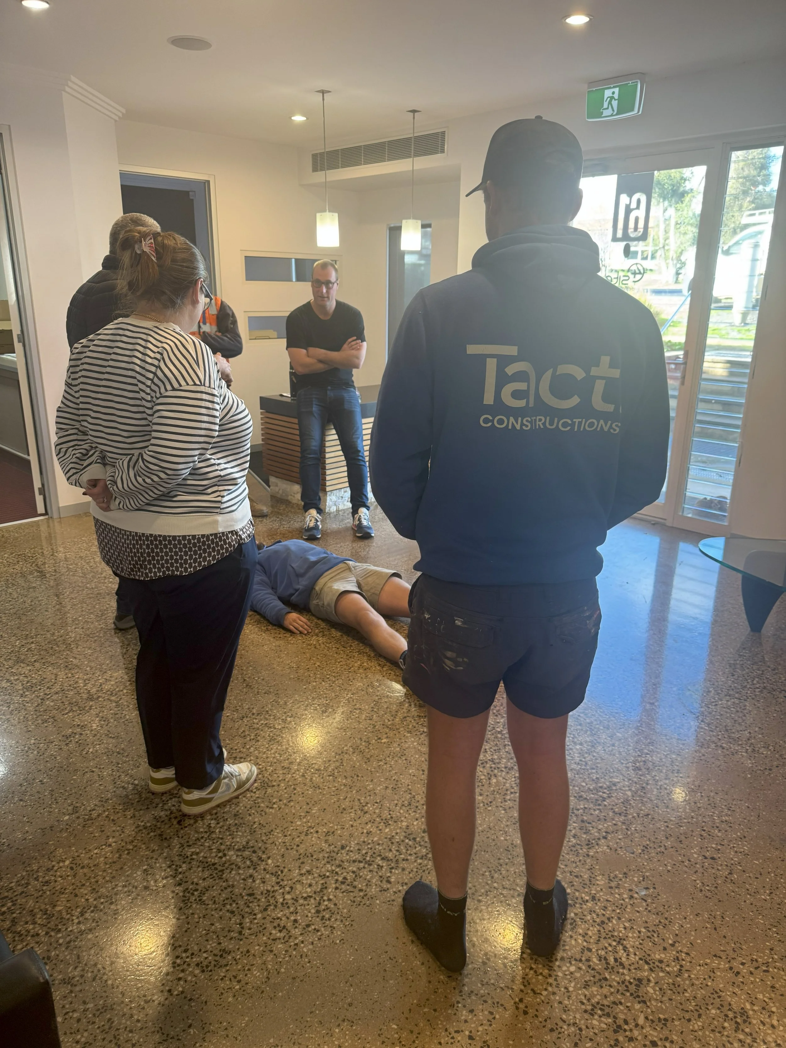 A man lies on the floor with four people standing around him inside a room, while another man in a hoodie with the words 'Tact Constructions' on the back watches.