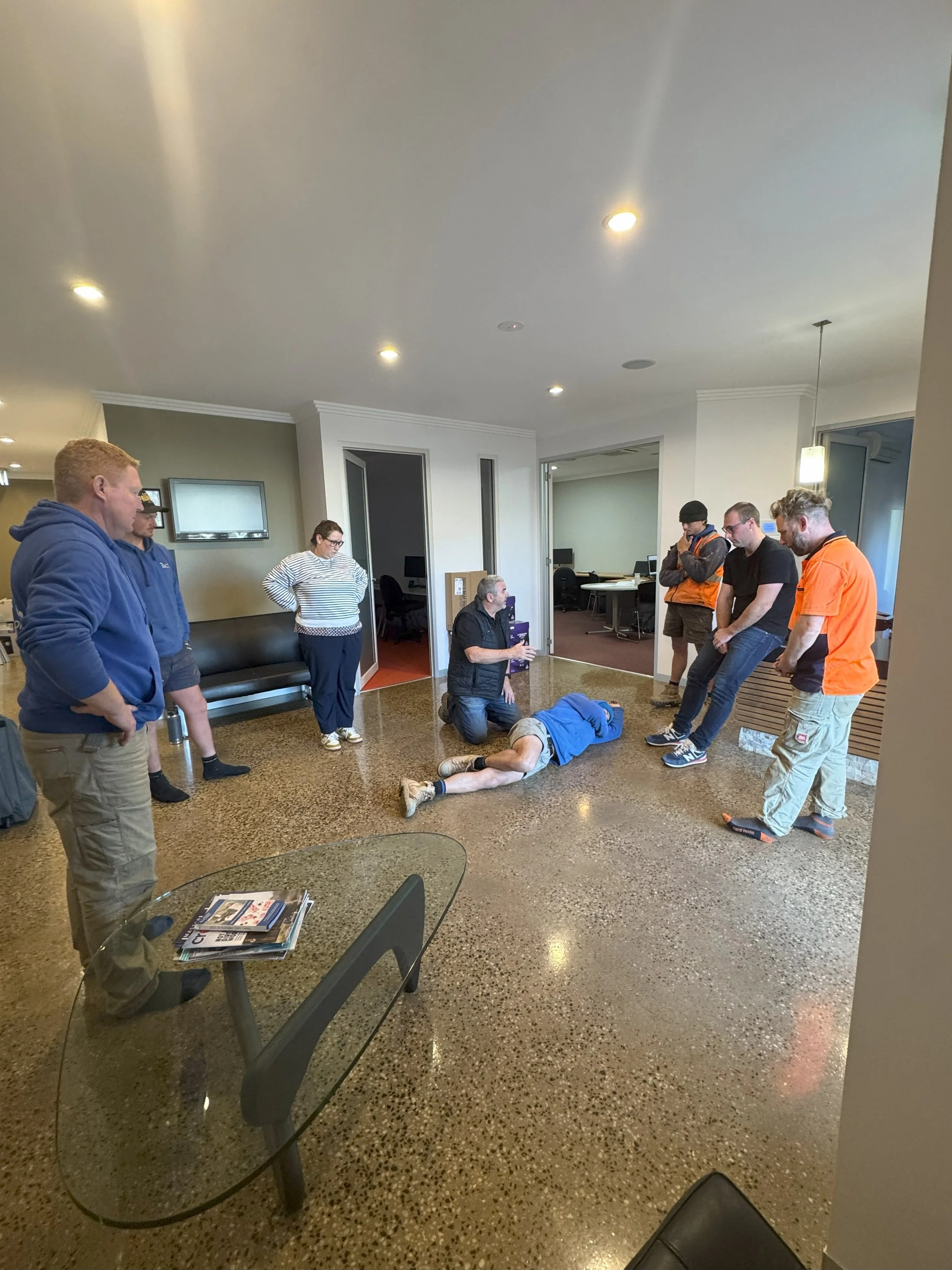 A group of people attending a CPR training session in an indoor space. A man is lying on the floor performing CPR while an instructor demonstrates. Others are watching. The room has a polished floor, a wall-mounted TV, and small tables.
