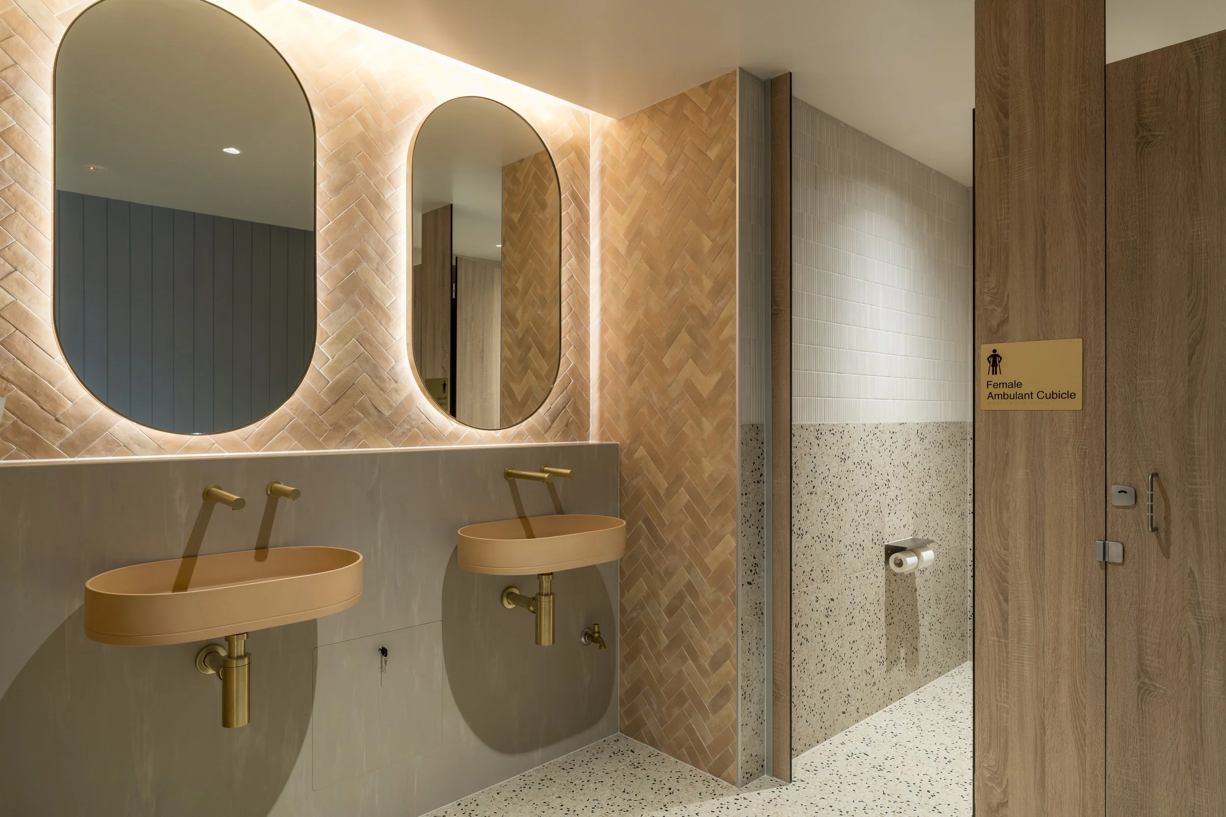 Public women's restroom with two beige sinks and three mirrors, wooden and tiled walls, terrazzo flooring, and a door labeled 'Female Ambulant Cubicle'.