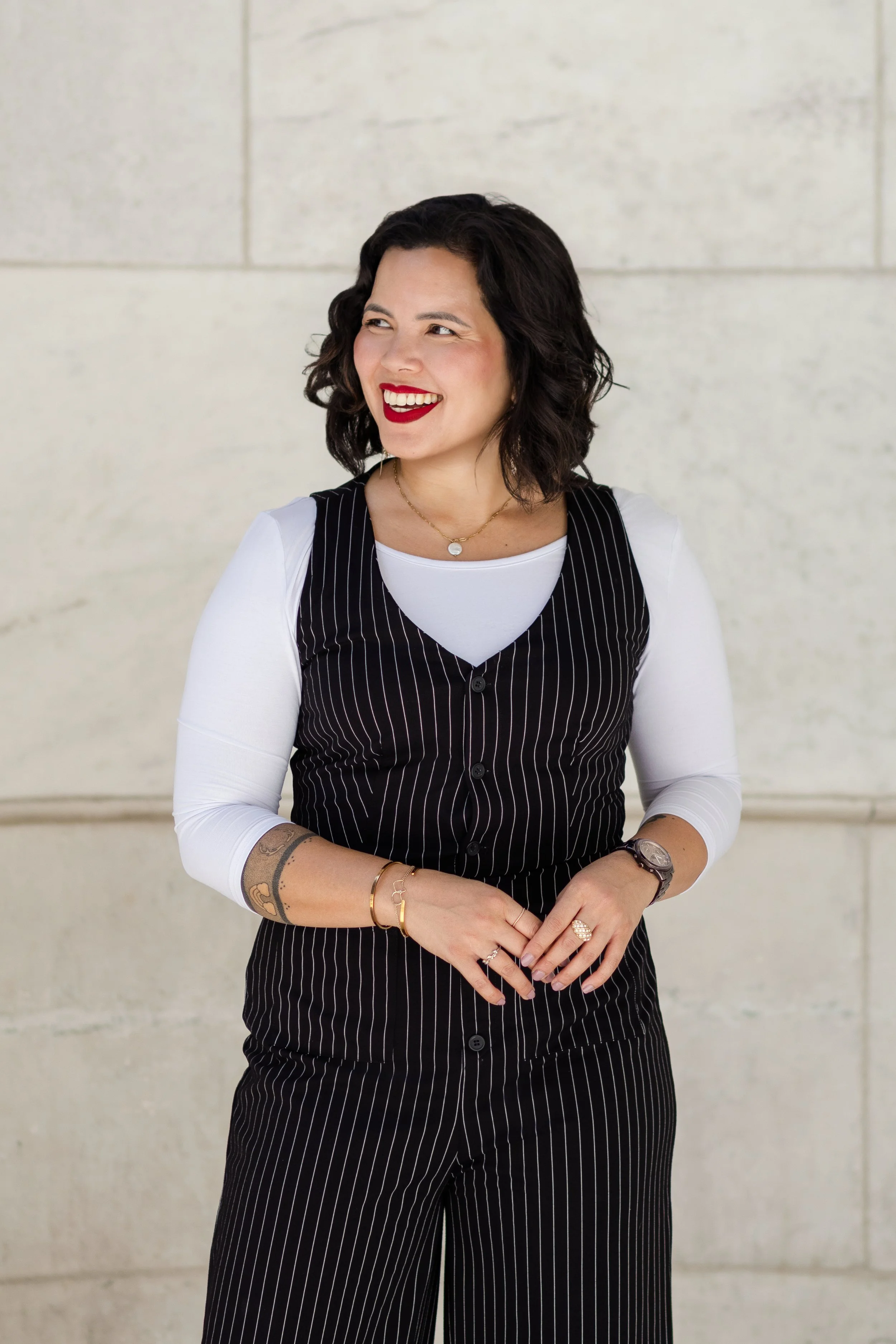A woman with wavy dark hair and red lipstick smiling and looking to her left, standing in front of a light-colored stone wall. She is wearing a black pinstripe vest over a white long-sleeve shirt, with various jewelry including rings, bracelets, a watch, and a necklace.
