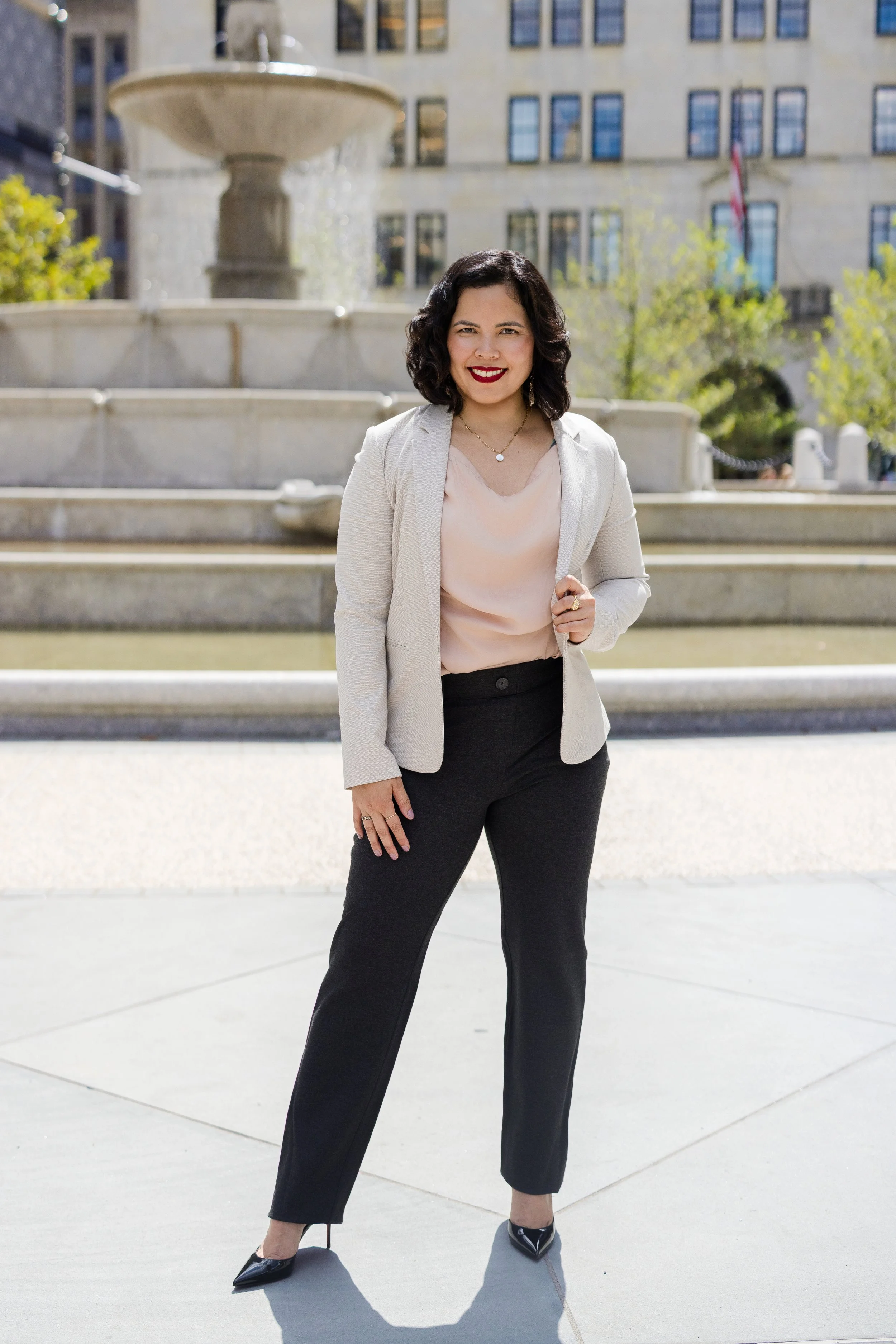 Woman with curly black hair, white blazer, pink blouse, black pants, and black high heels standing outdoors in front of a stone fountain and a building with blue window frames.
