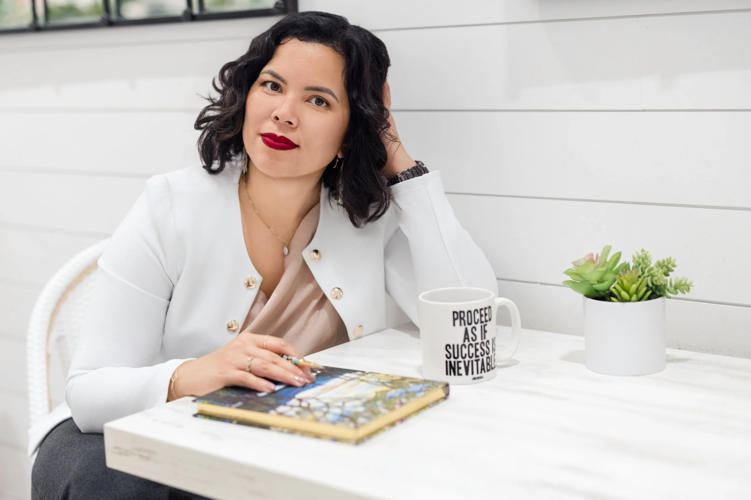 A woman with black curly hair and red lipstick sitting at a white table, holding a book, with a white mug and green succulent plant on the table. The mug has black text that reads "PROCEED AS IF SUCCESS IS INEVITABLE." She is wearing a white blazer with gold buttons and a beige top.