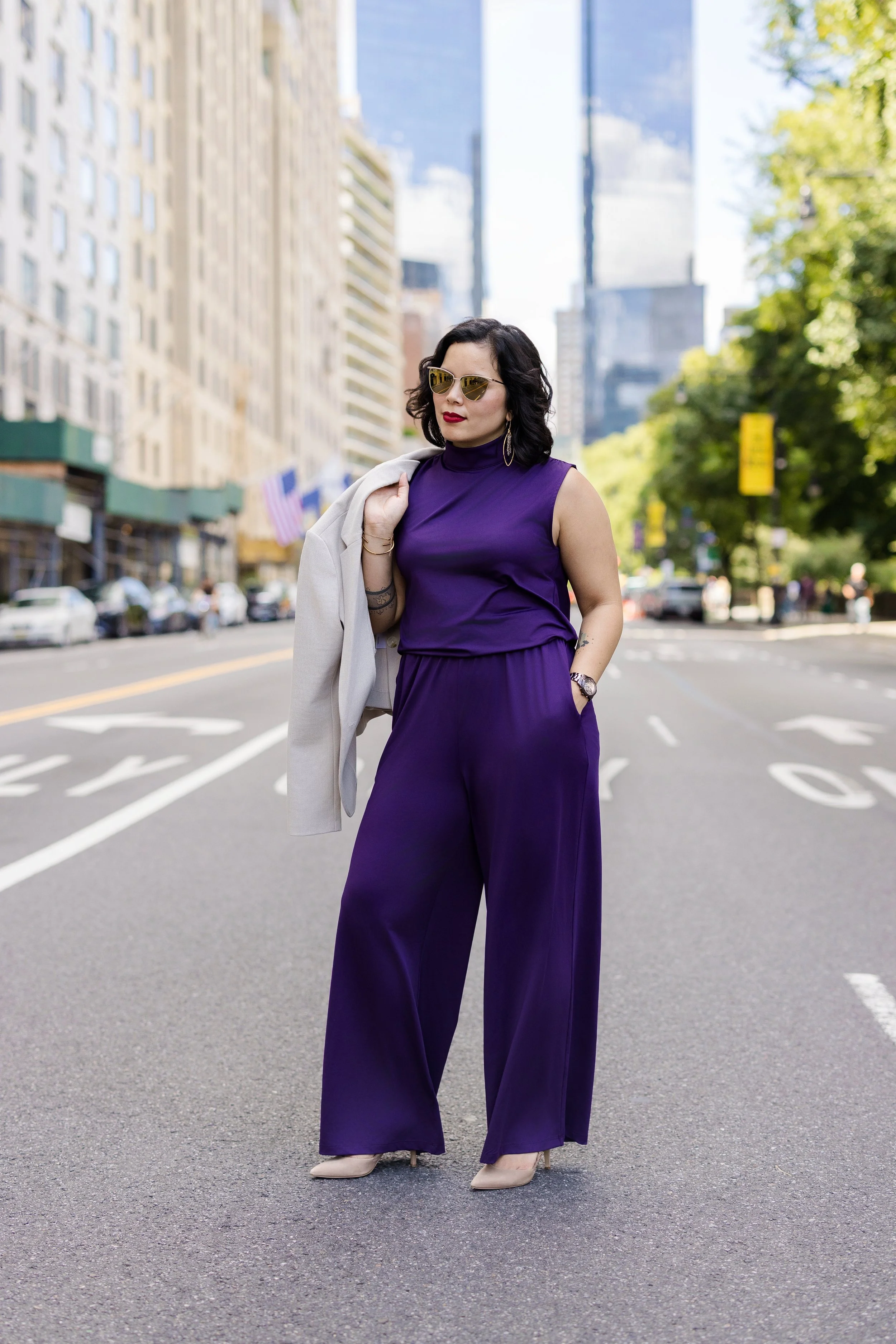 A woman in a purple jumpsuit with a white blazer draped over her shoulder, wearing beige high heels, sunglasses, and red lipstick, standing on a city street with tall buildings and trees in the background.
