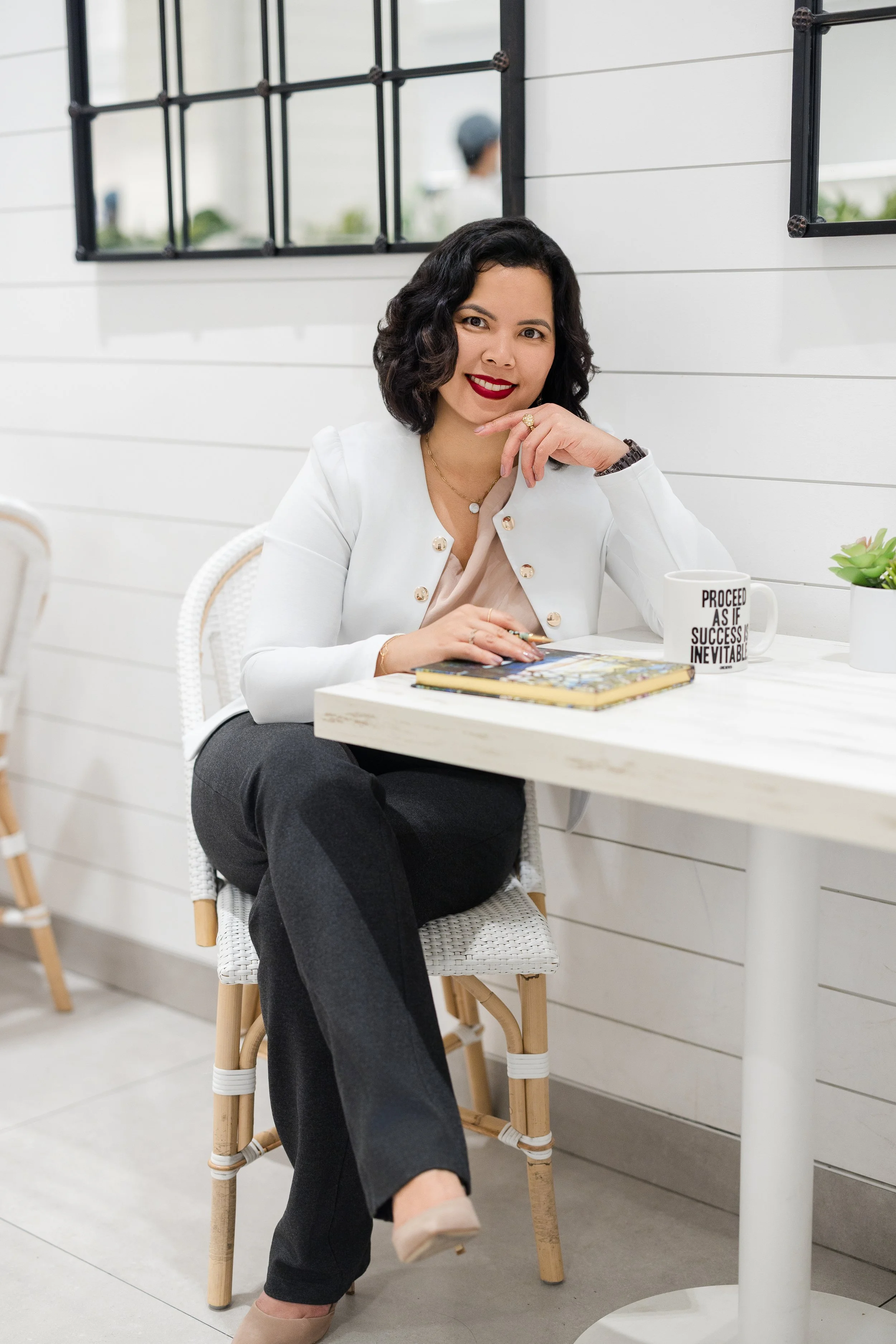 A woman with dark wavy hair and red lipstick sitting at a white table in a bright room with white shiplap walls. She is wearing a white blazer with gold buttons and gray pants. She has a gold necklace, a ring, and a watch. There is a white mug with black text, a small potted succulent, and a book on the table. Behind her is a large mirror with black metal gridwork, reflecting a blurred figure in the background.