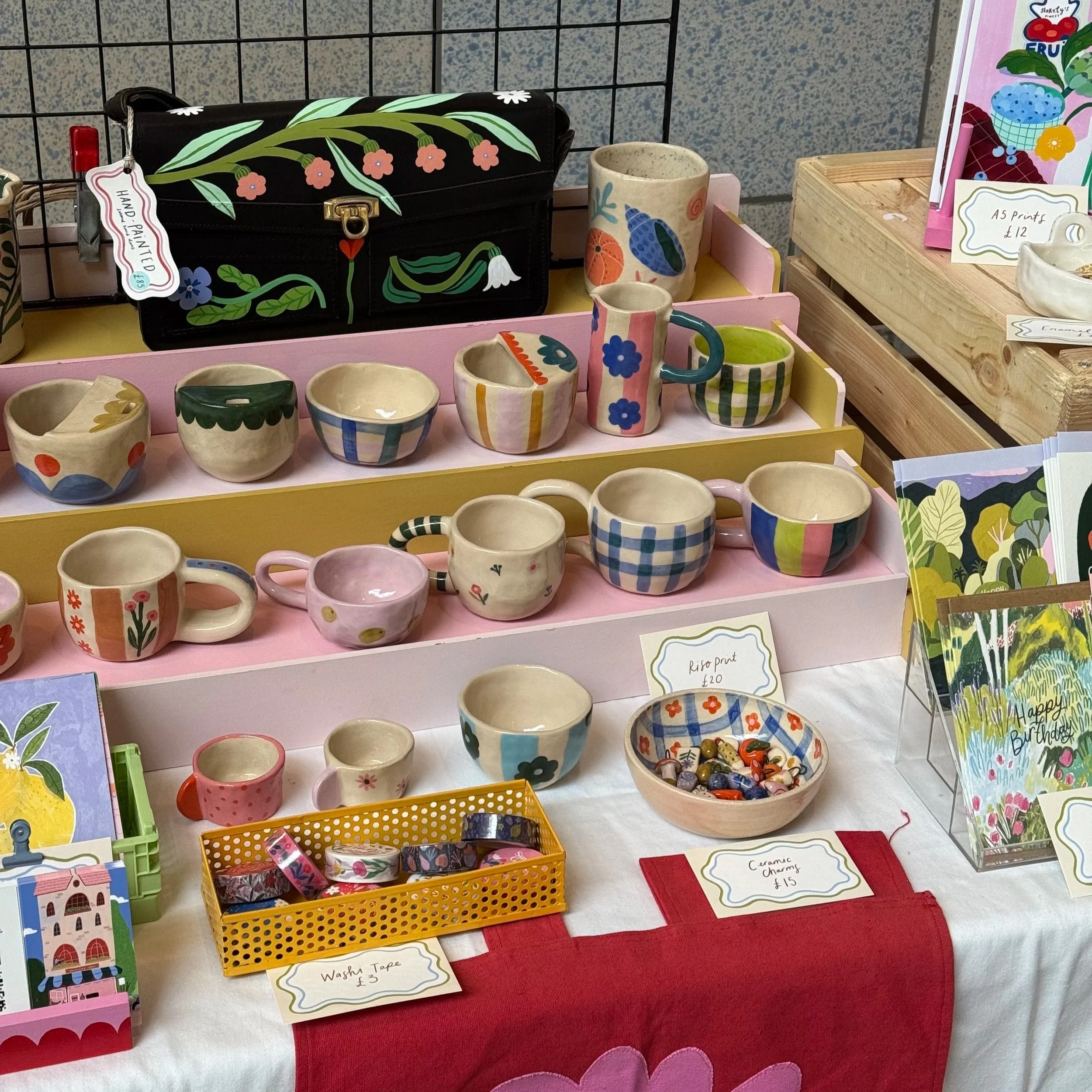 Colorful handmade ceramic cups and bowls displayed on shelves. Some cups have floral, checkered, or striped patterns. A black bag with floral designs is seen in the background. Small signs with prices are visible, indicating items such as wash tape and ceramic charms. Various cards and decorative items are on the table.