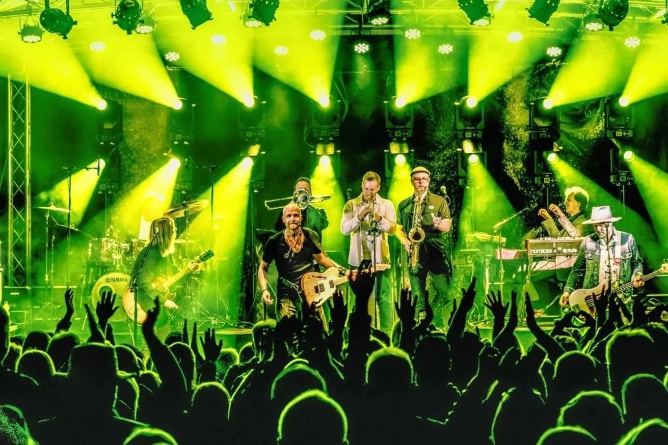 Eläytyi lavalla bändi, jonka jäsenet soittavat instrumentteja ja ovat valaistuja kirkkaan keltaisilla valoilla, yleisöä juhlitaan edessä.