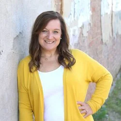 A woman wearing a yellow cardigan and white top, smiling while leaning against a textured wall.