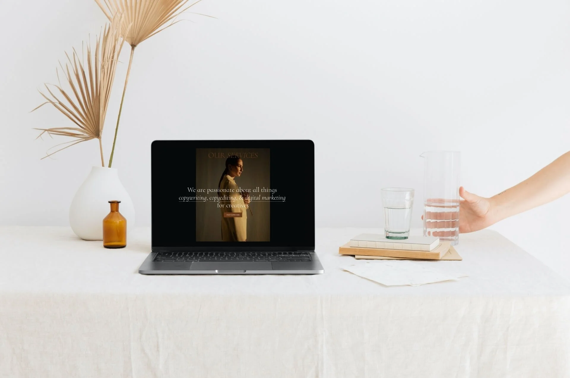 A minimalist workspace with a laptop displaying a webpage about copywriting and digital marketing. Beside the laptop, there's a vase with dried palm leaves, a small brown bottle, a stack of books, and a glass of water. A hand is reaching for a carafe of water on a light-colored tablecloth.