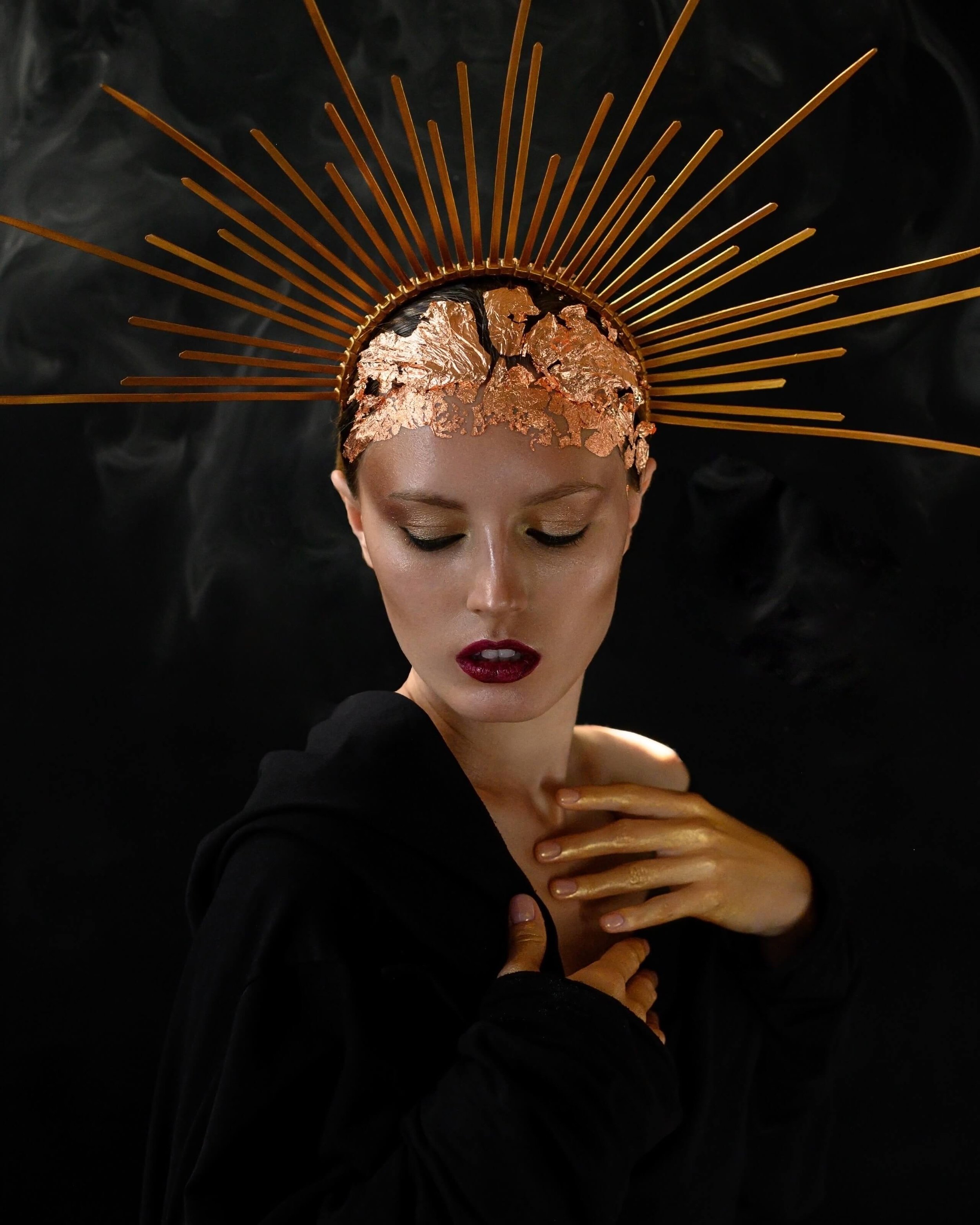 Woman wearing a golden spiked headdress with closed eyes and dark makeup against a dark background.