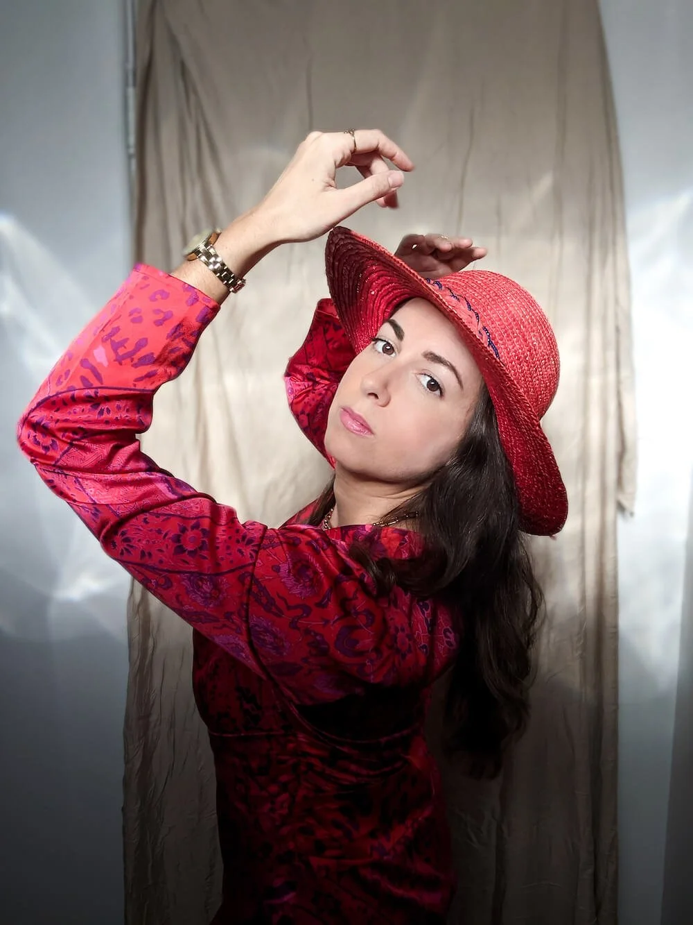 A woman in a red floral dress and wide-brimmed red hat poses with her hand raised, looking directly at the camera. The background consists of beige fabric.
