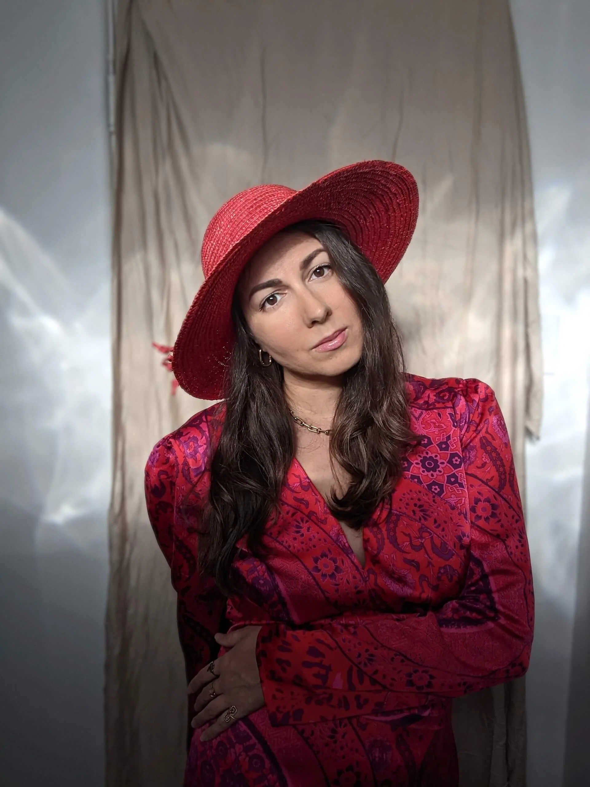 A woman with long dark hair wearing a large red sunhat and a red patterned dress, posing in front of a beige curtain background.
