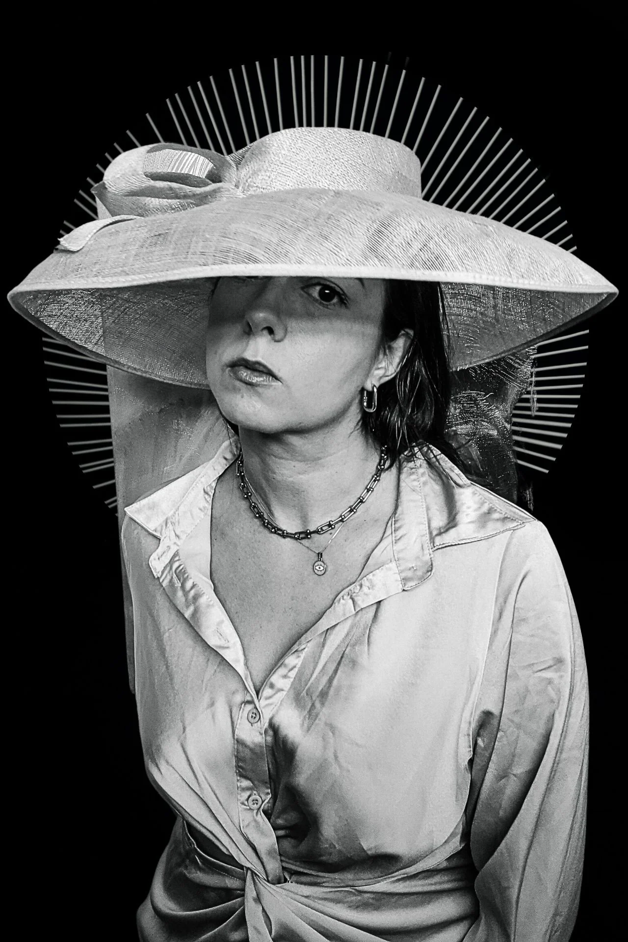 Black and white photo of a woman wearing a wide-brimmed hat, a button-up shirt, and layered necklaces, against a dark background.