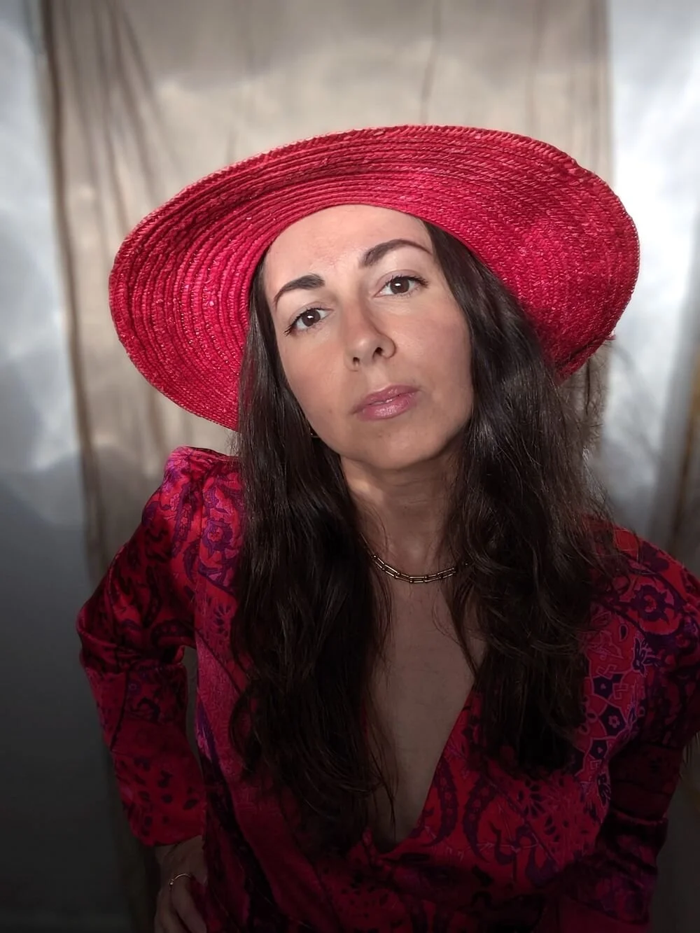 Woman with long dark hair wearing a large red sunhat and a red patterned dress, looking at the camera.
