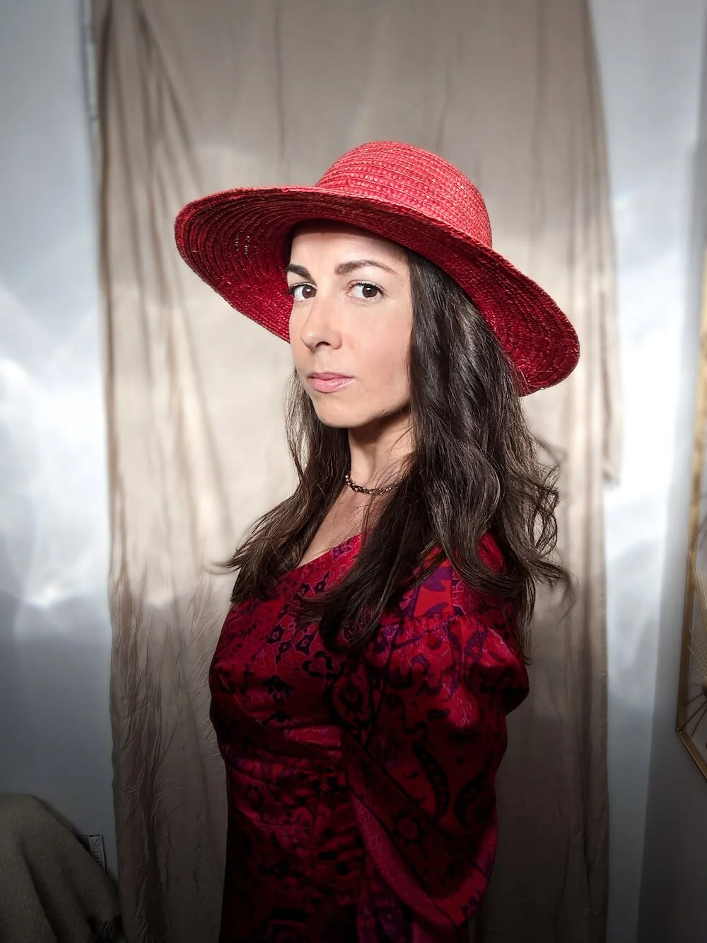 A woman with long dark hair wearing a red wide-brimmed hat and a red patterned dress standing indoors in front of a beige curtain.