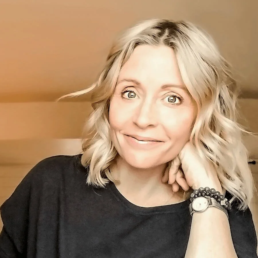 Woman with blonde hair smiling, wearing a black shirt and a watch, with her hand resting on her chin.
