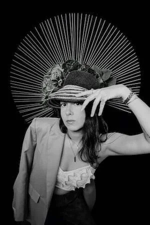 A woman wearing a hat decorated with flowers and fabric, posing with her hand near her face in front of a circular, radiating background.