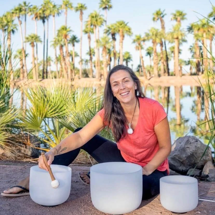 A woman playing crystal singing bowls by a lake with palm trees in the background.