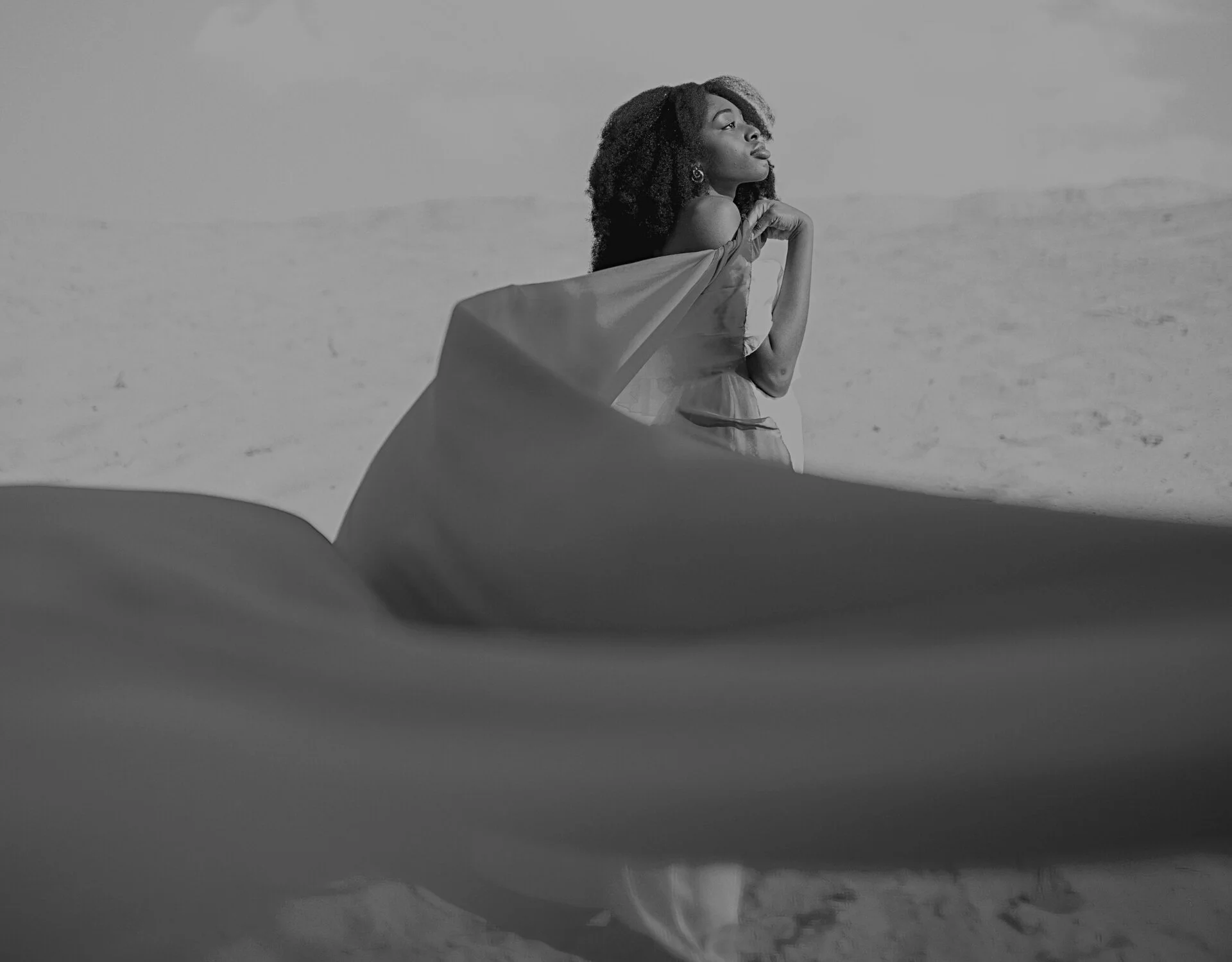 A woman with curly hair stands in a desert landscape, looking into the distance, with a large fabric piece draped over her shoulder and a flowing dress.