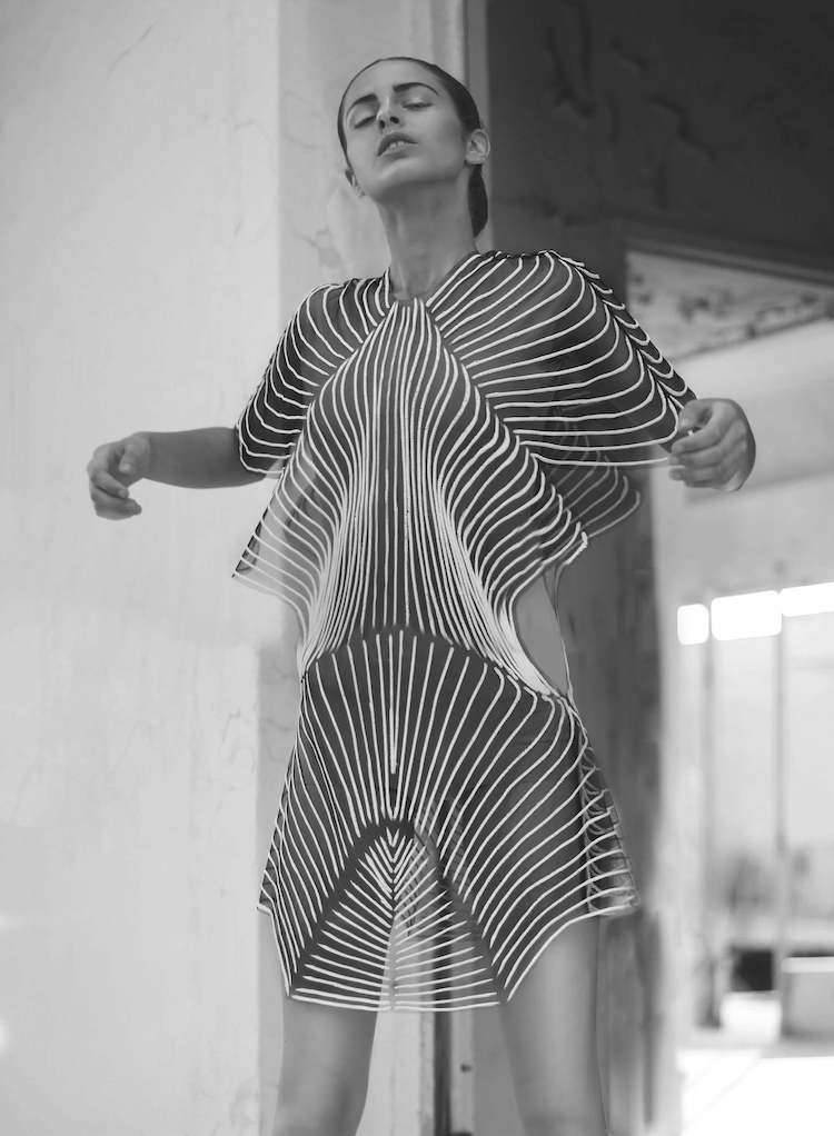 A woman in a striped dress poses with her eyes closed, standing indoors near a wall with a window in the background.